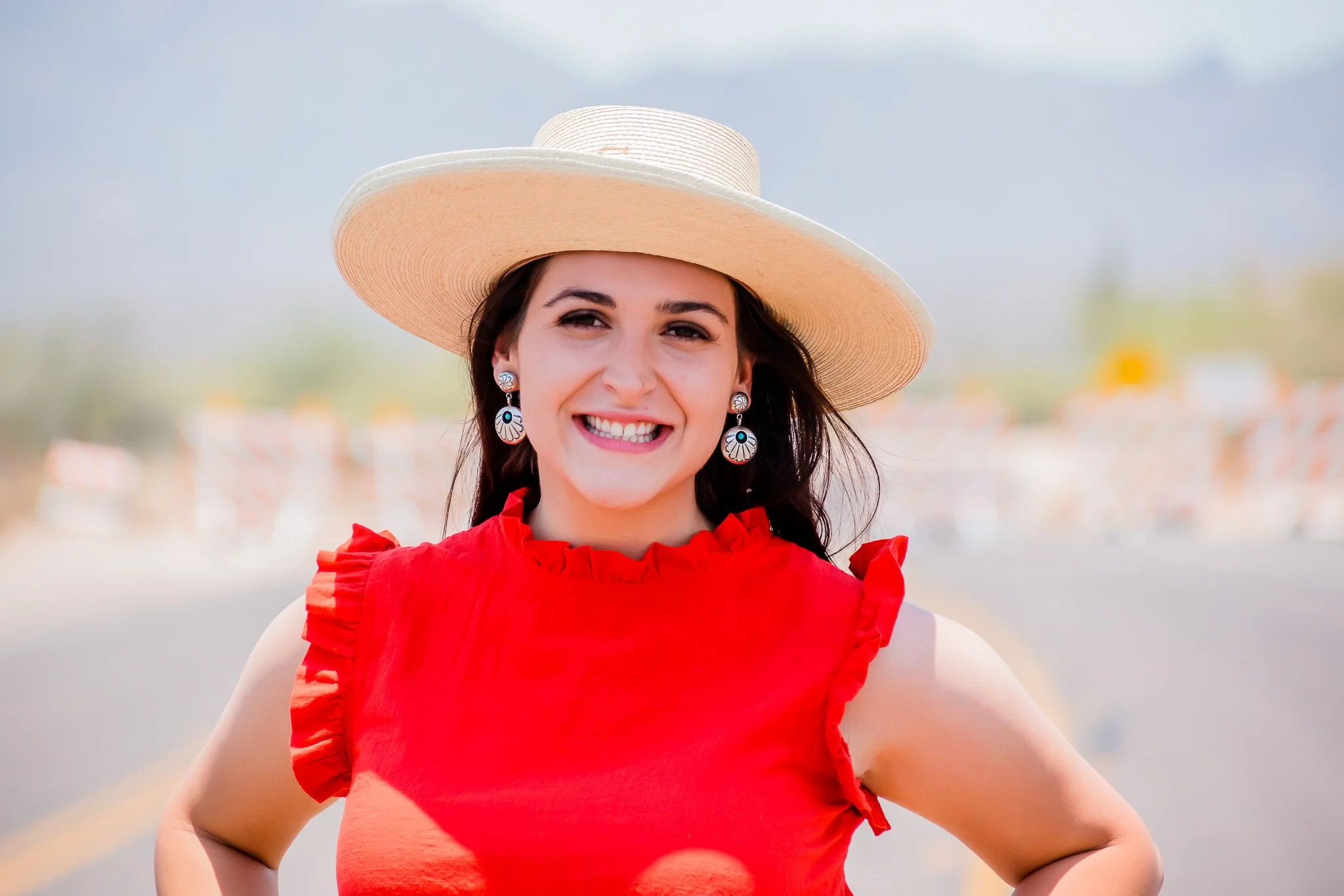 A smiling young woman wearing a wide-brimmed hat, red ruffled top, and earrings, standing outdoors on a sunny day.