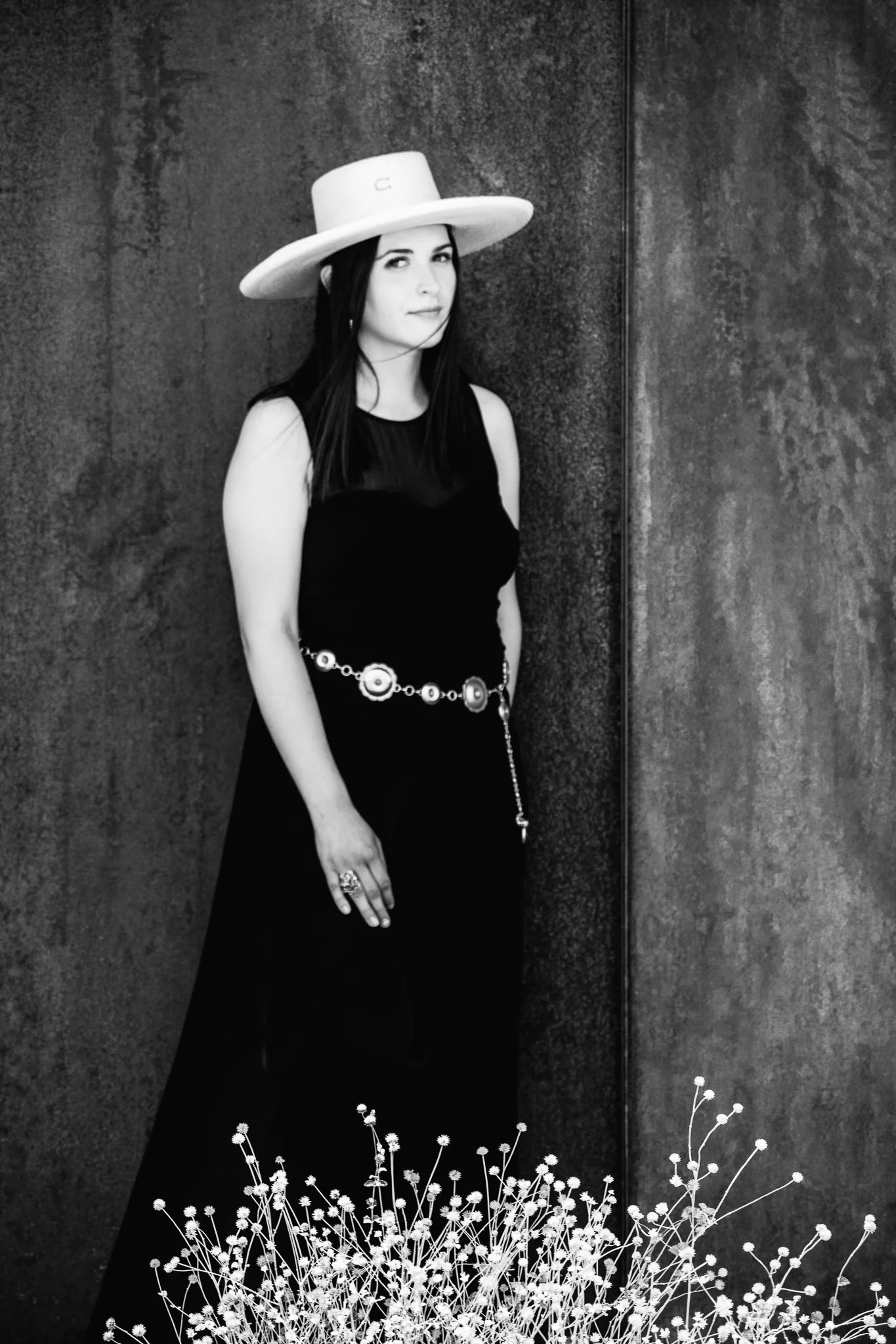 Black and white photo of a woman wearing a wide-brimmed hat, a sleeveless black dress, a decorative belt, and rings on her fingers, standing against a textured wall with plants in the foreground.