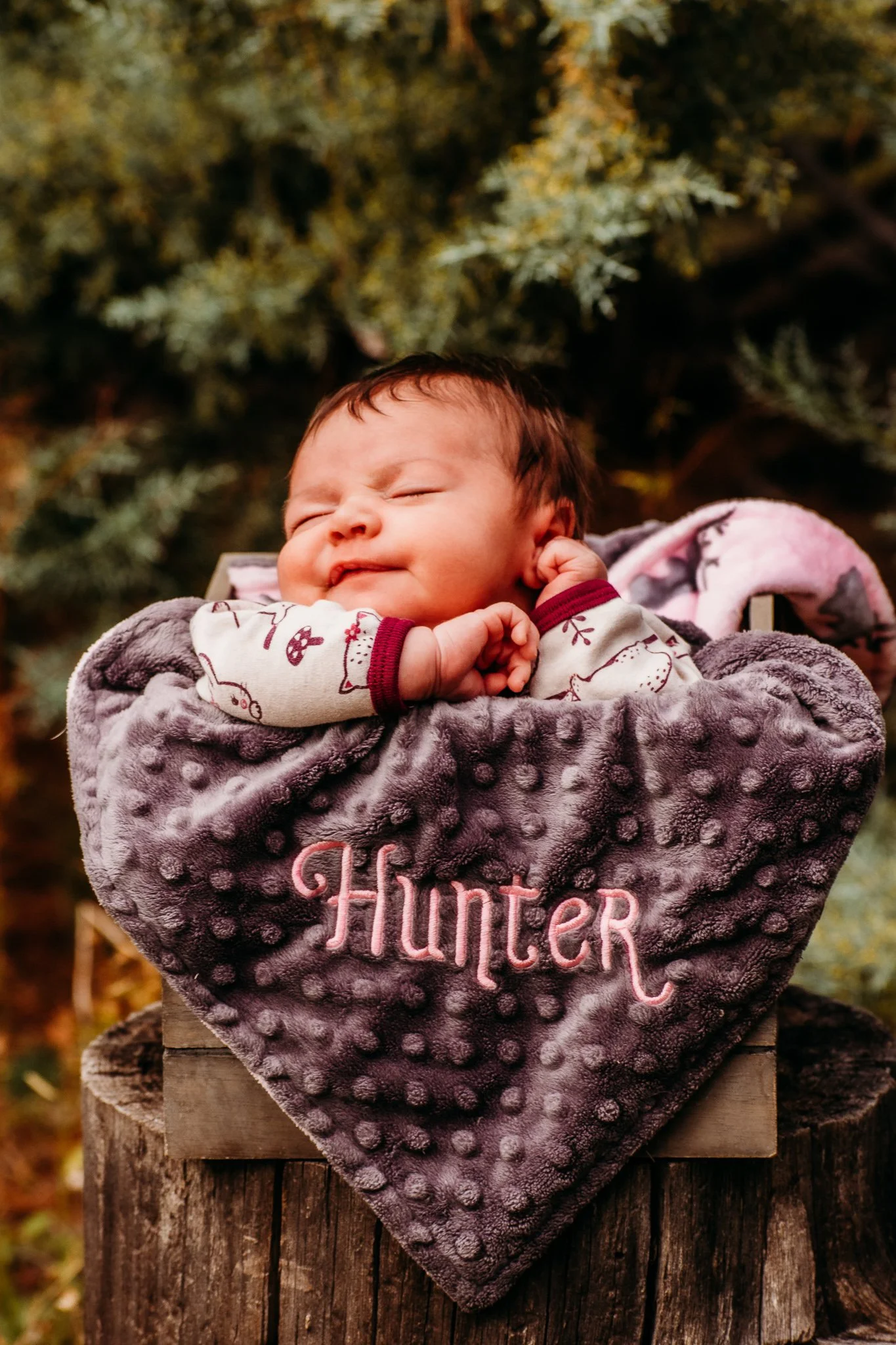 A sleeping baby nestled in a soft purple blanket with pink embroidered letters spelling 'Hunter'. The baby is outdoors with a background of greenery.