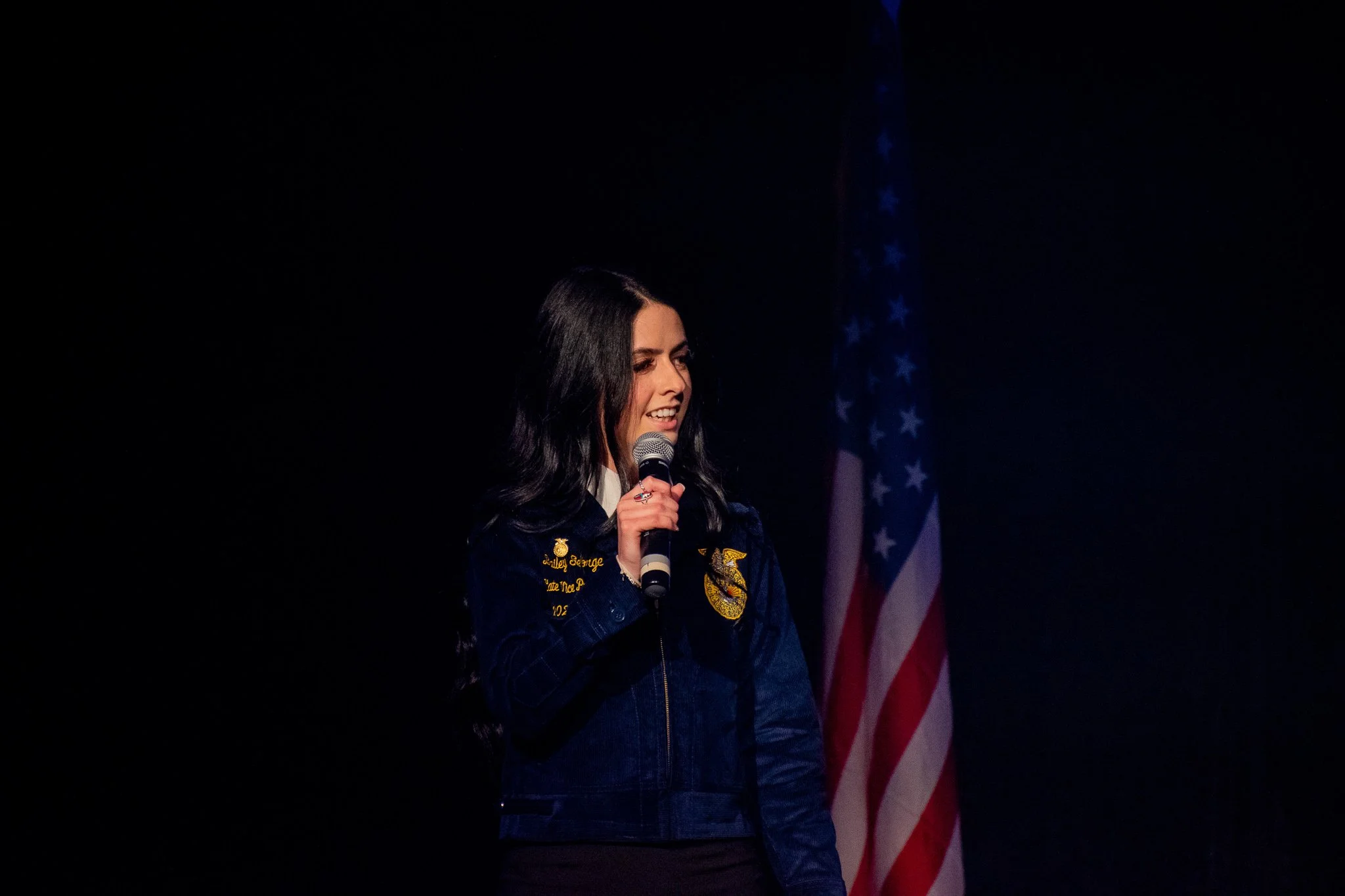 A woman with long black hair speaking into a microphone on stage, wearing a dark blue jacket with embroidered patches, and waving an American flag in the background.