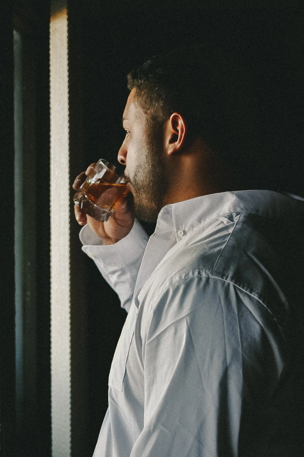 A man in a white shirt drinking from a glass of whiskey while standing against a dark background.