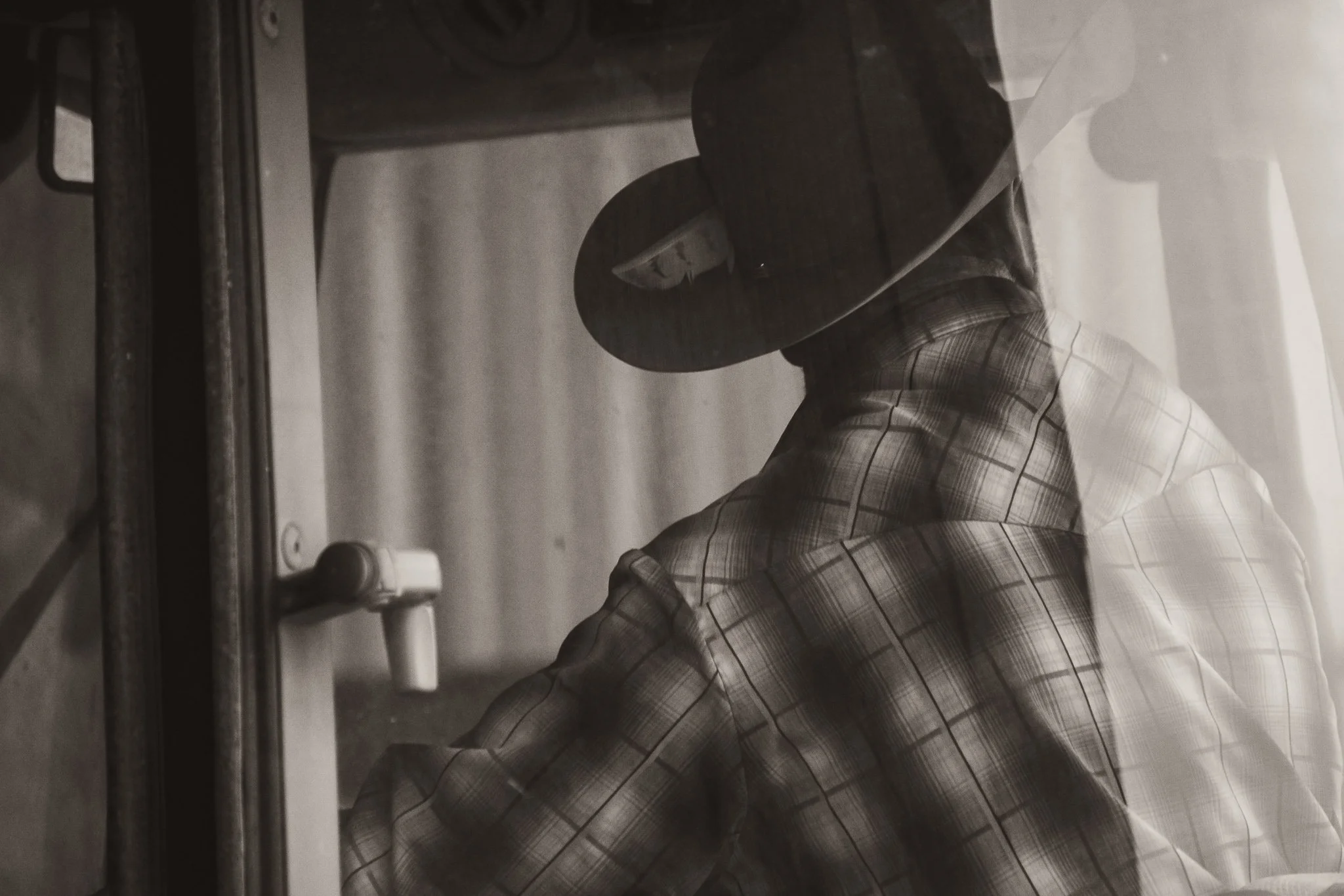 A person wearing a plaid shirt and wide-brimmed hat, leaning forward and facing a window or glass surface, reflected in the glass.