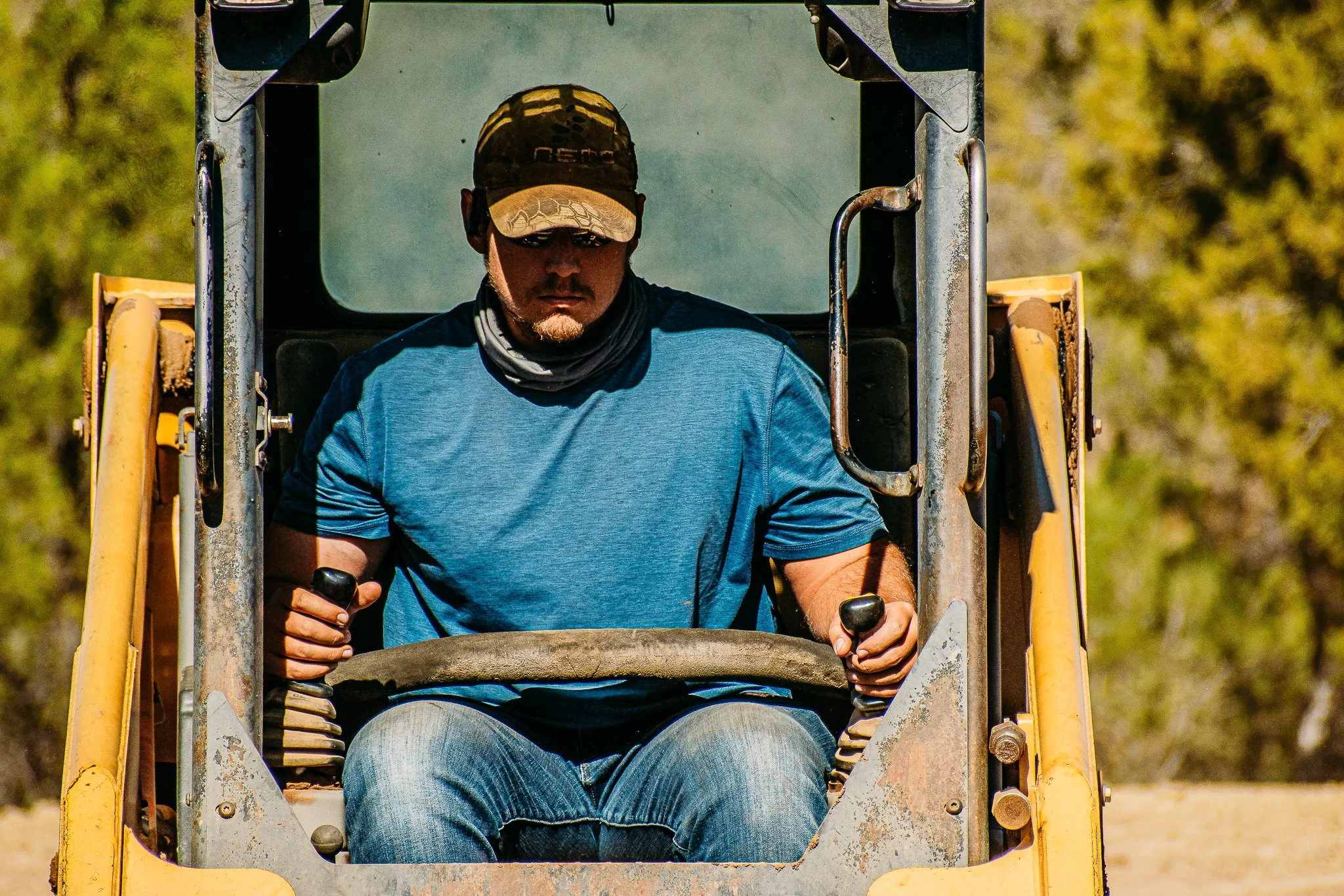 A man operating heavy construction equipment, sitting in the cabin and looking down with control levers in each hand, outdoors with blurry green trees in the background.