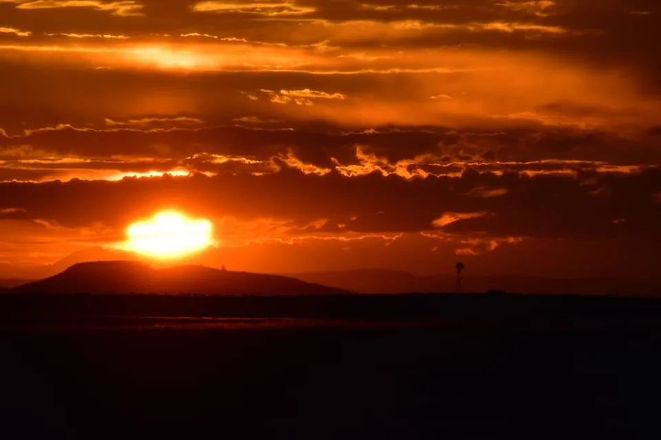 Sunset over a desert landscape with a silhouette of a hill and cloud-filled sky.