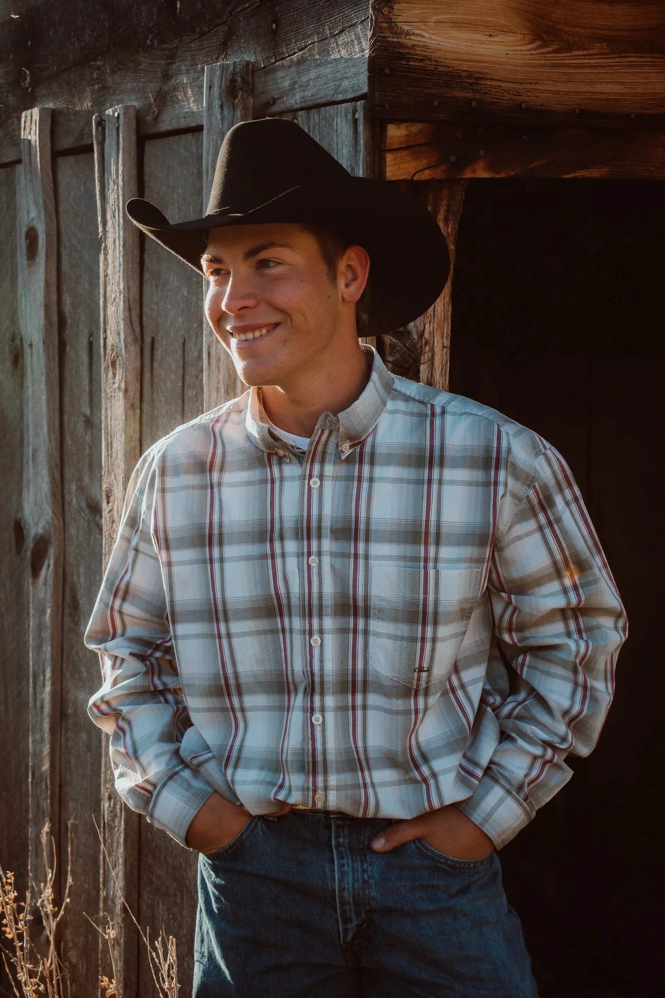 A young man wearing a cowboy hat, plaid shirt, and jeans standing outdoors in front of a rustic wooden building, smiling and looking to the side.