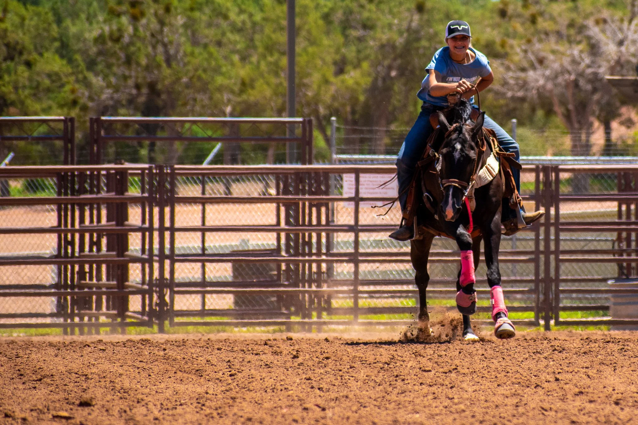 A young girl riding a galloping black horse on a dirt arena, wearing a blue shirt, helmet, and pink leg wraps, with a smile on her face.