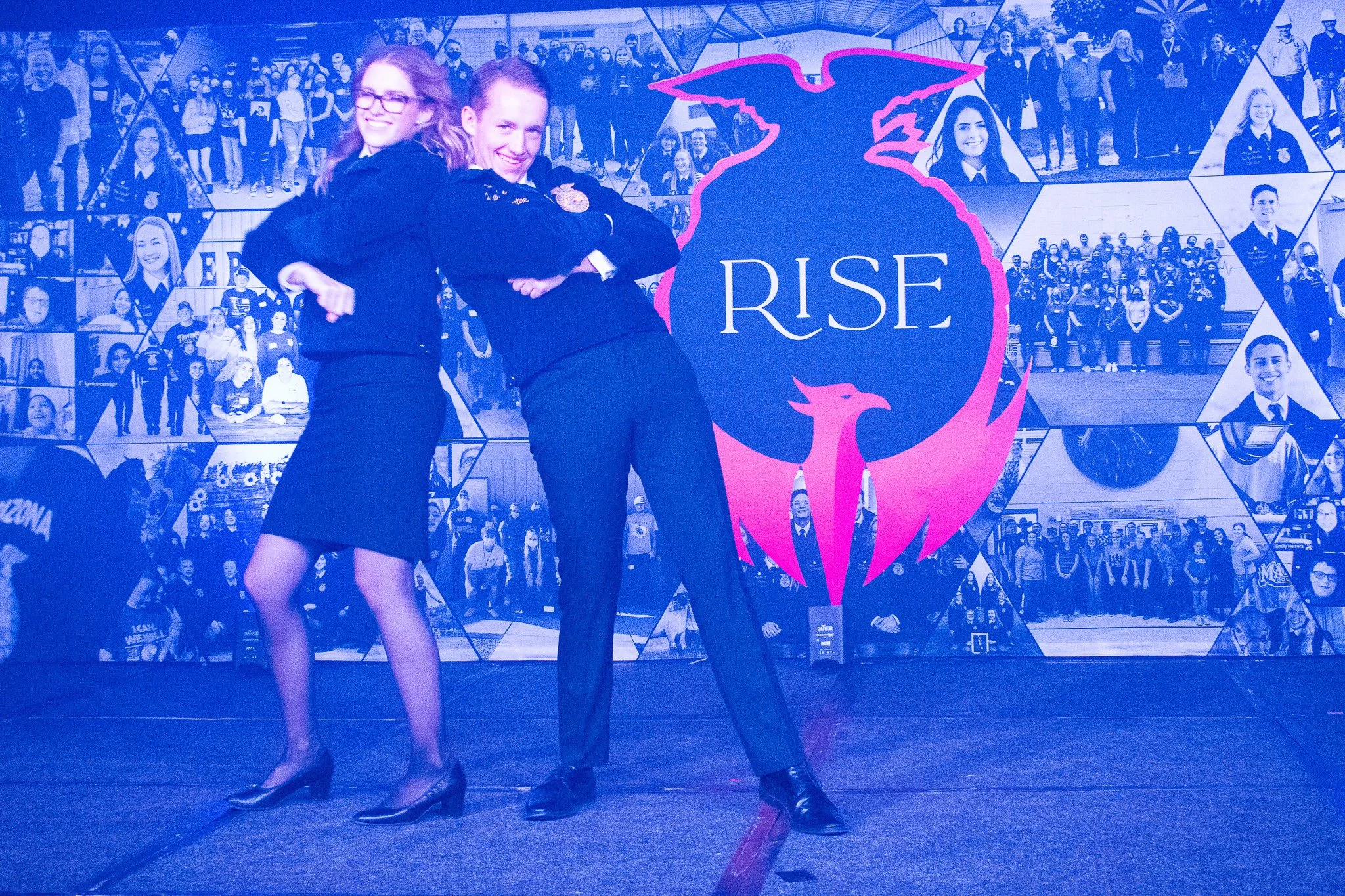 Two students in official uniforms posing confidently in front of a large banner with a blue and pink emblem reading 'RISE' and a bird graphic, with a collage of school photos in the background.