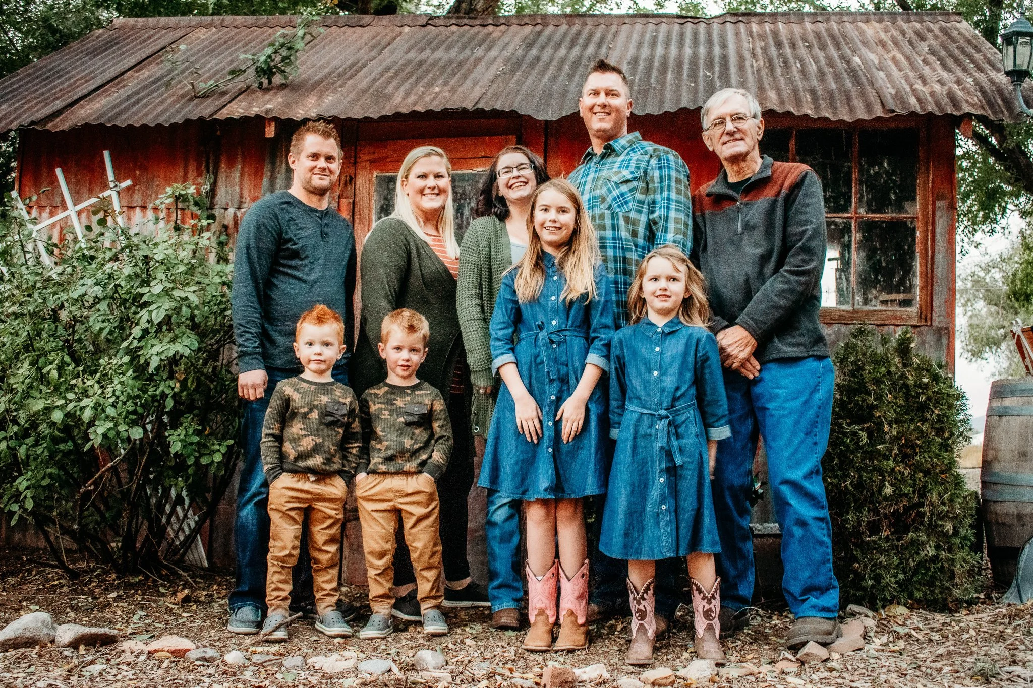 A multi-generational family of nine stands in front of a rustic wooden shed, outdoors, smiling at the camera. The group includes children, parents, and grandparents, dressed in casual and western-style clothing.