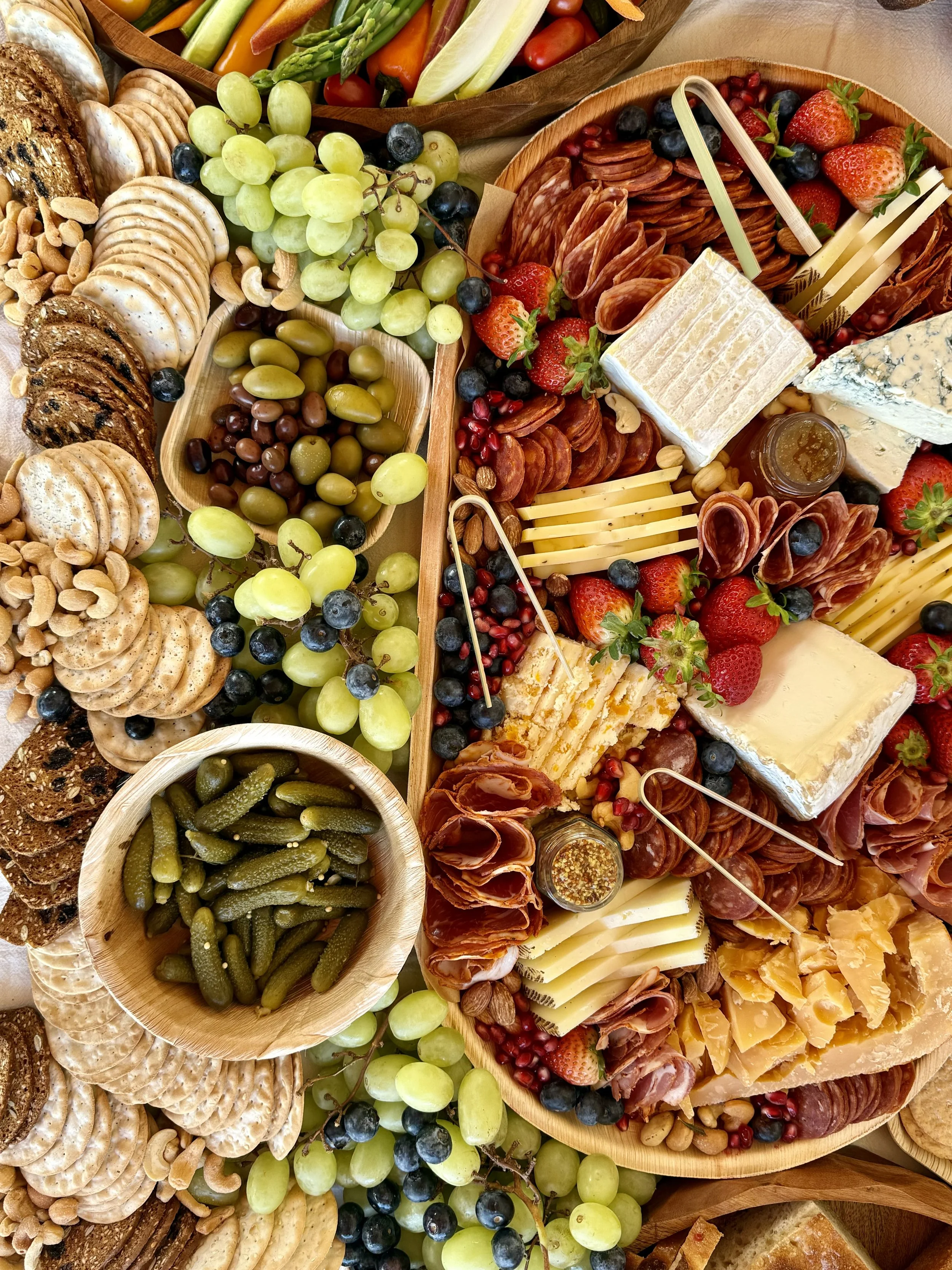 Charcuterie board with assorted cheeses, cured meats, fresh grapes, cherry tomatoes, cucumber slices, a jam jar, and crackers.