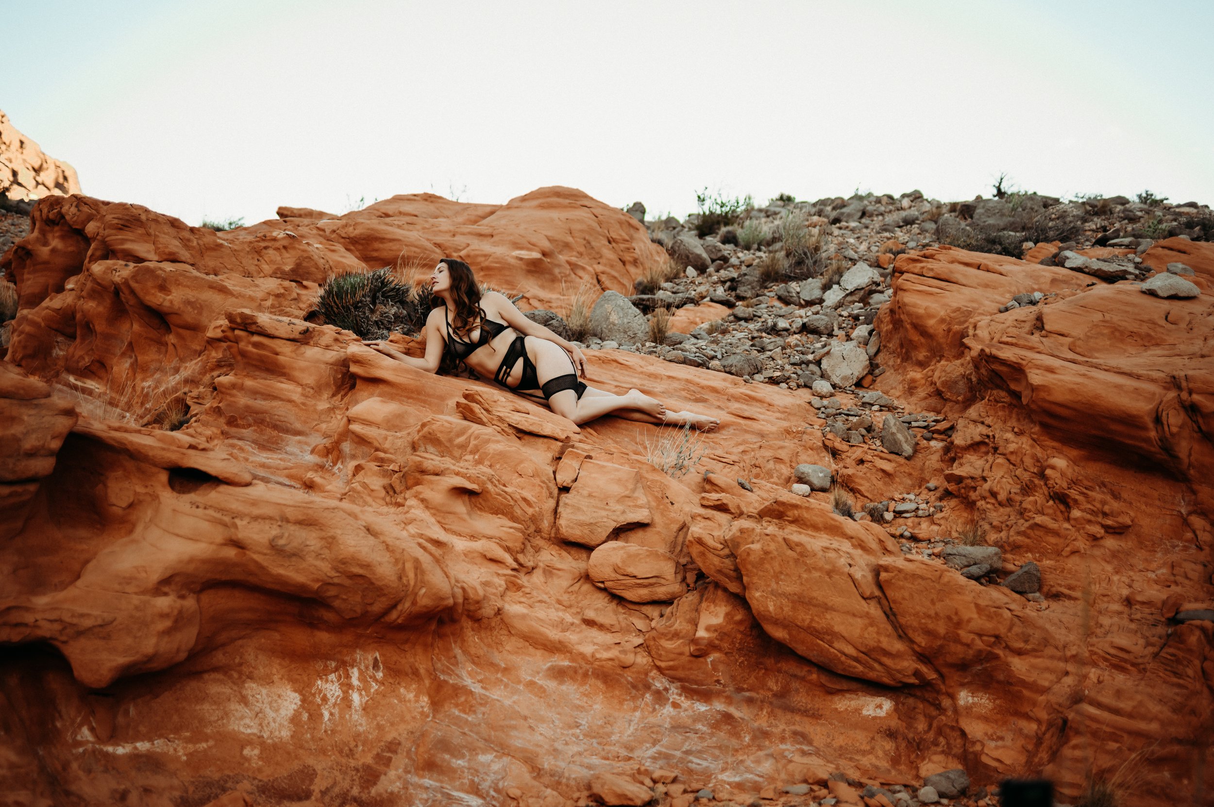 A woman with tattoos, wearing black clothing and high heels, walks on a dirt path in a desert landscape with sparse vegetation, abandoned car, and mountain range in the background.