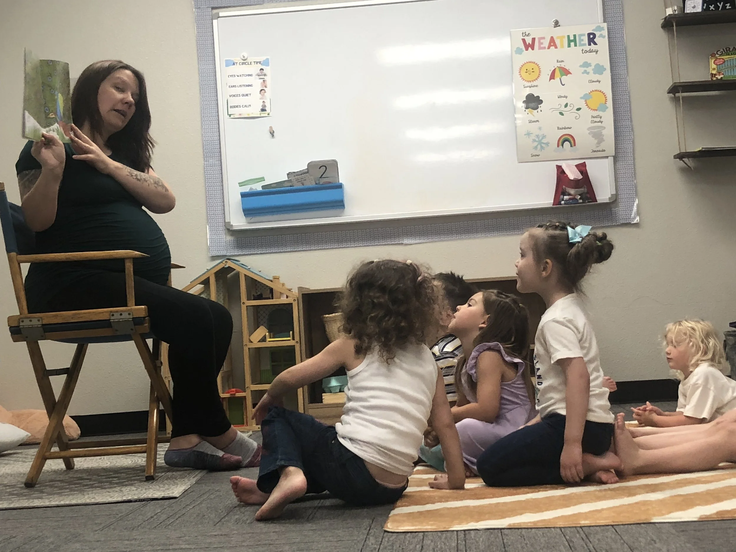 A teacher sitting on a chair in a classroom, reading a book to a group of children sitting on a rug on the floor, with a whiteboard and colorful weather chart behind them.