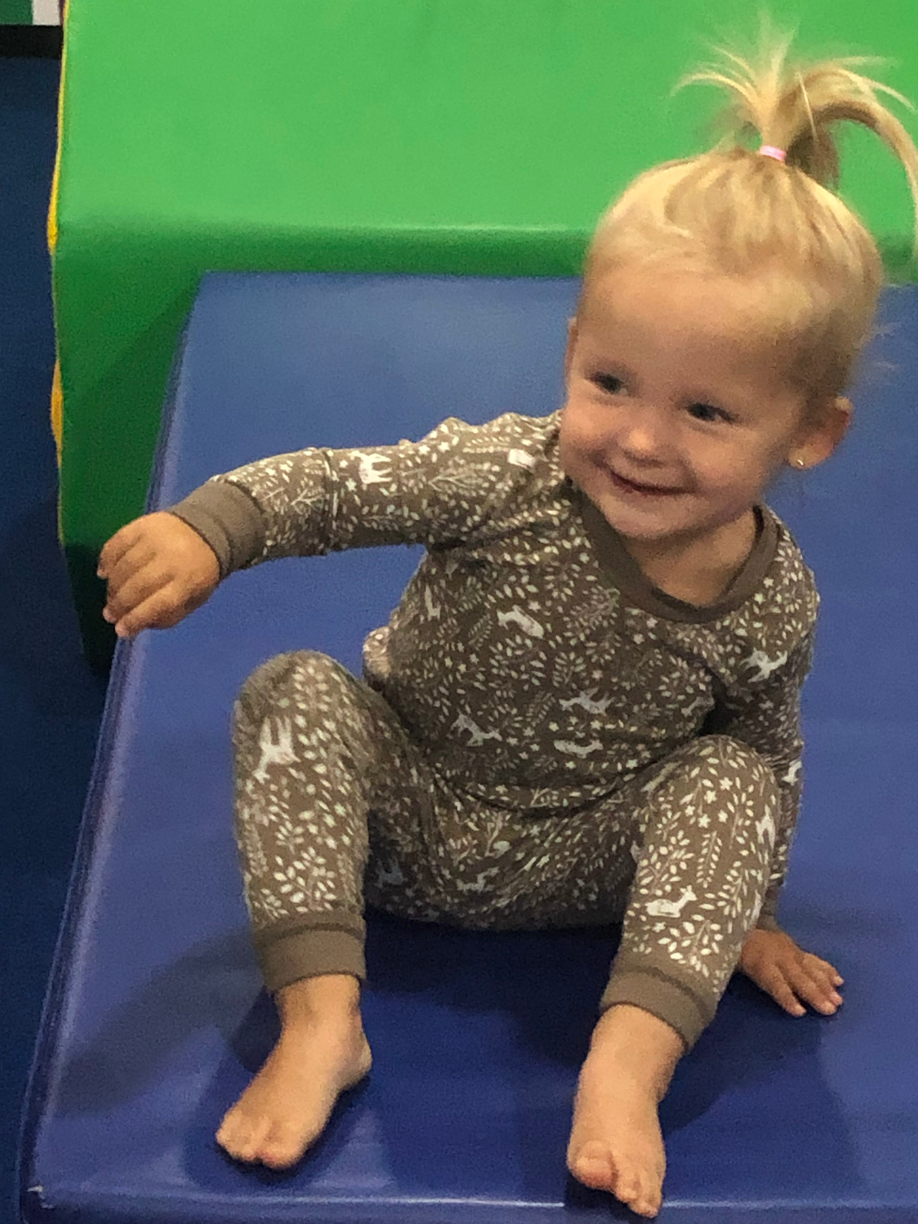 A young girl with blonde hair tied in a pink hair tie, sitting happily on a blue mat in an indoor play area with green and blue padded walls, wearing patterned pajamas with white animals and foliage design.