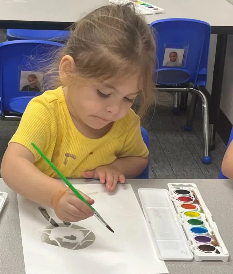 A young girl in a yellow shirt painting with watercolors at a table in a classroom.