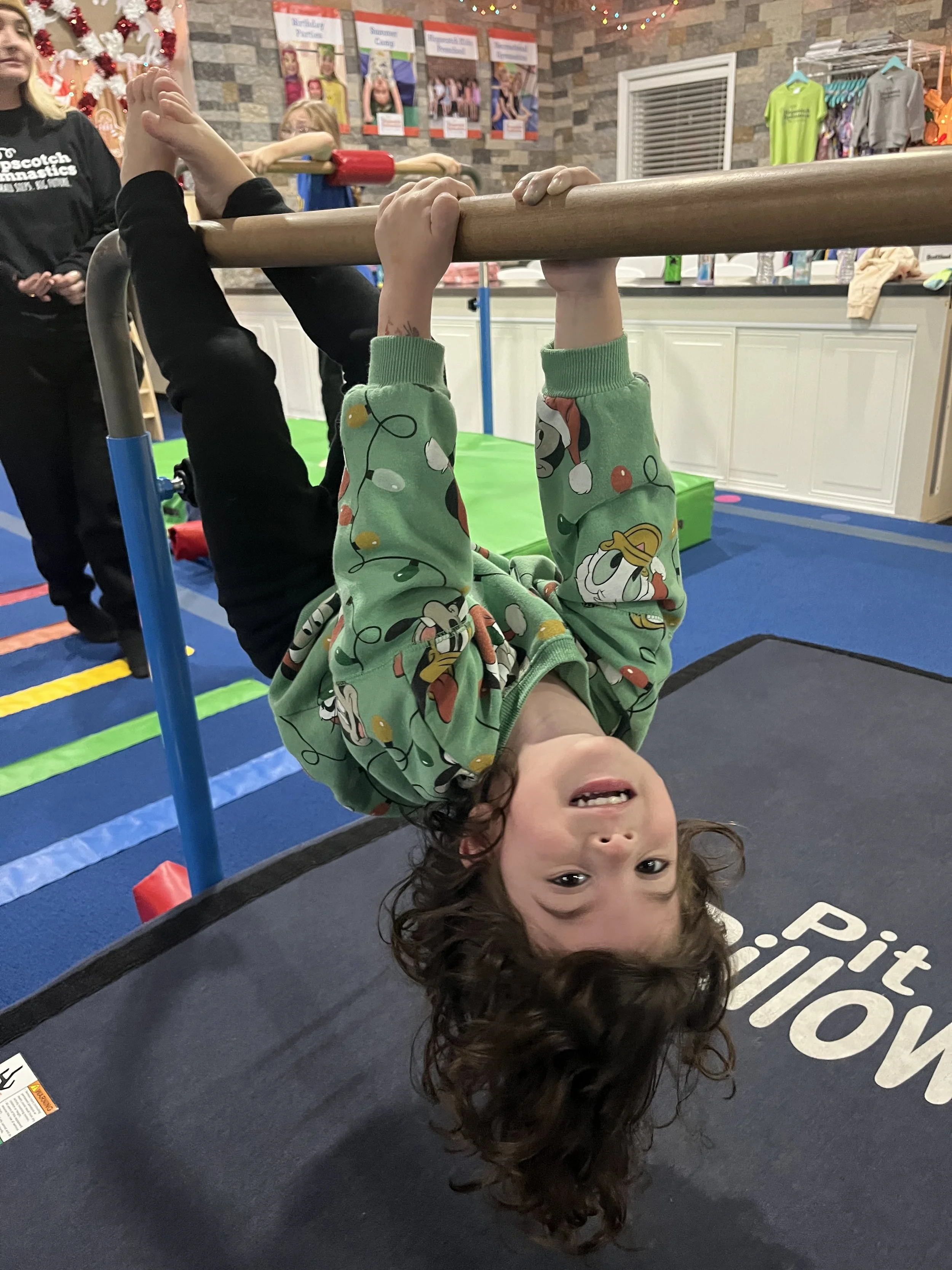 Children playing in a gymnastics gym, crawling through a tent-like structure made of purple mats on the gym floor.
