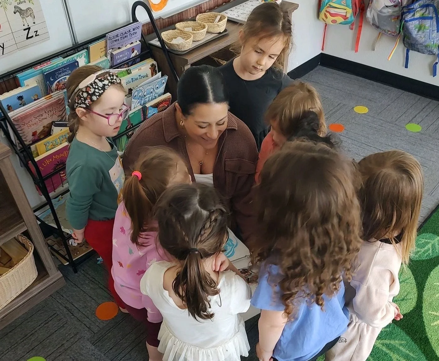 A teacher surrounded by young students gathering closely around her in a classroom. The students are looking at something in the teacher's hands. The classroom has colorful decorations and a carpet with colored dots.