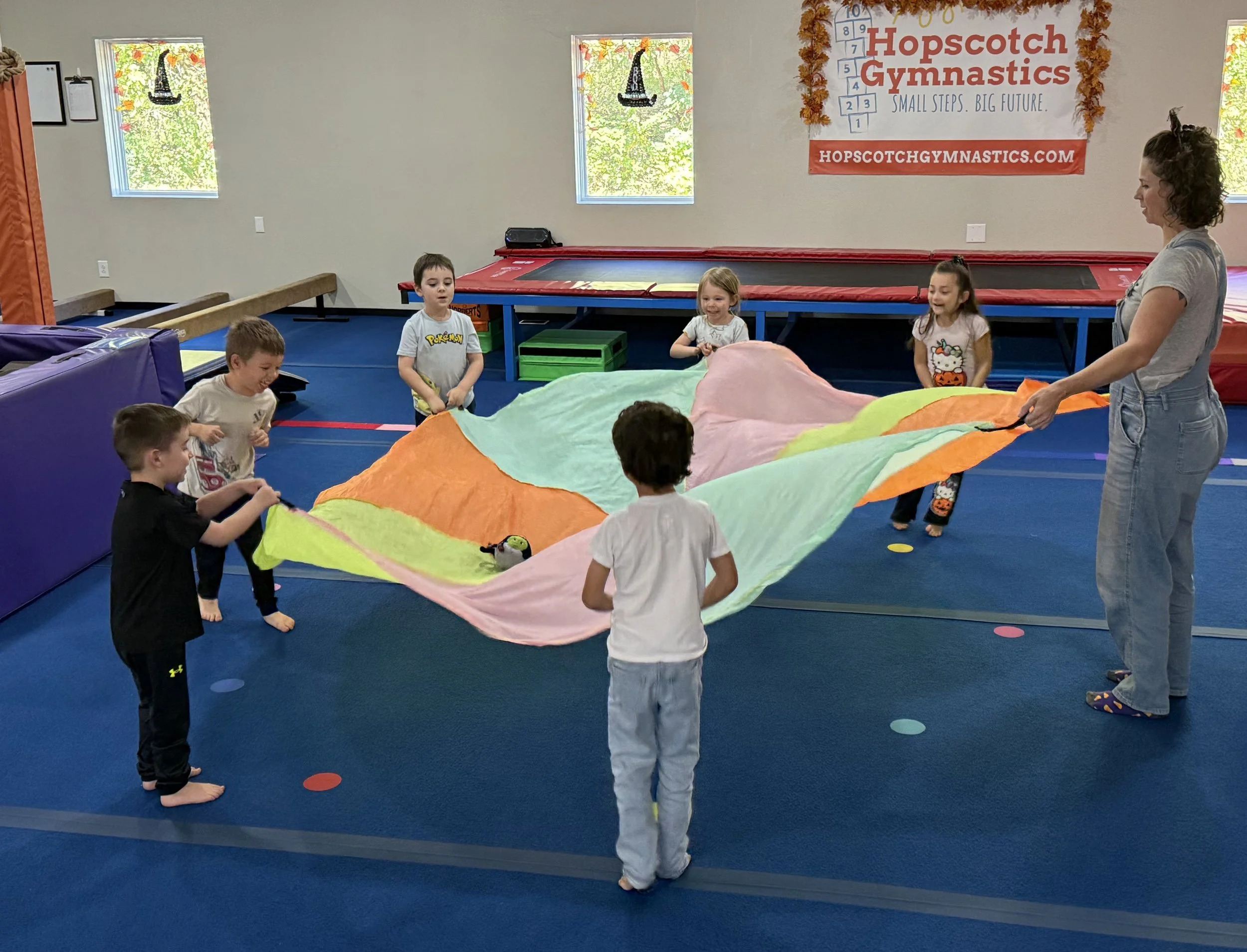 Children and an adult playing with a colorful parachute in a gymnastics class. Logo in the background reads 'Hopscotch Gymnastics.'