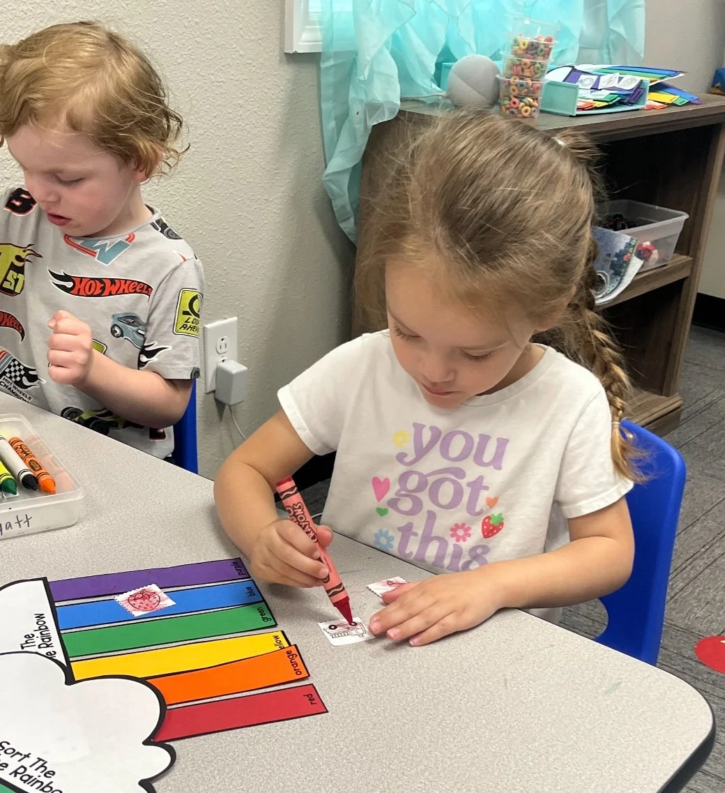 A young girl is coloring valentines at a desk in a classroom. A boy with curly red hair and cartoons on his shirt is sitting nearby.