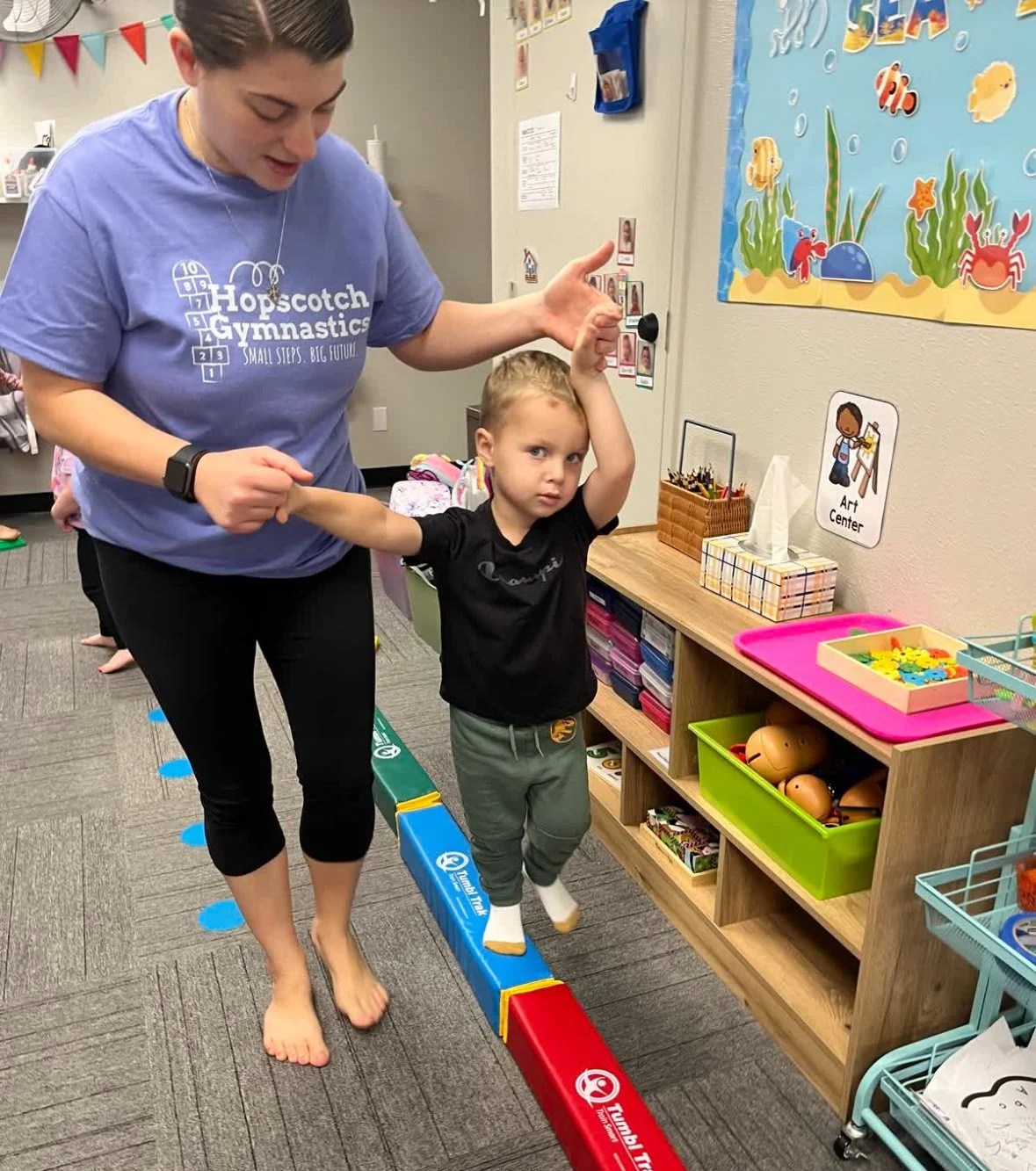 A young boy in gray sweatpants and a black shirt balances on a colorful balance beam while holding hands with an adult woman in a blue T-shirt and black pants, assisting him in a playroom.