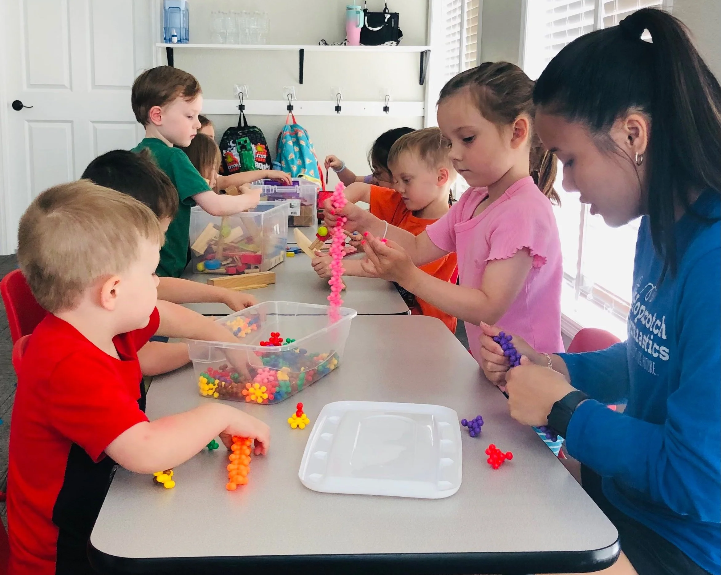 Children and a coach sitting at a table making bead crafts, with colorful beads and bead creations on the table and in containers.