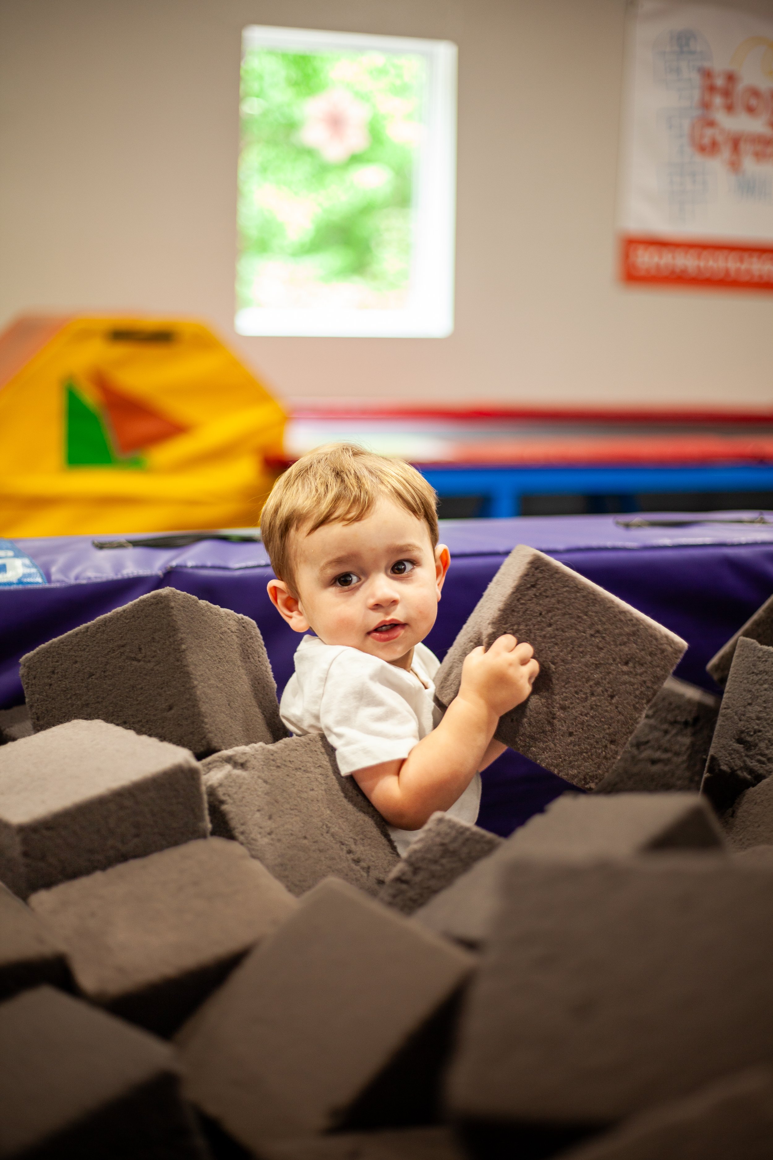 Children playing in a gymnastics gym, crawling through a tent-like structure made of purple mats on the gym floor.