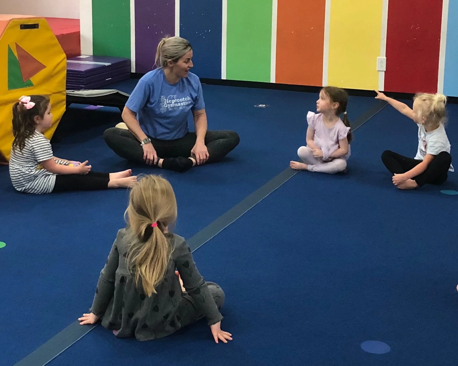 A woman leading a gymnastics class with four young girls sitting on a blue mat in a colorful gymnasium.
