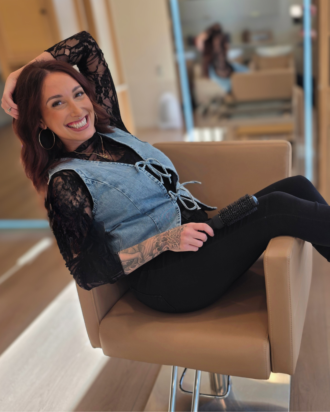 A woman with reddish-brown hair, tattoos on her arm, wearing hoop earrings, a black lace top under a denim vest, smiling and sitting in a salon chair with her legs up, holding a hairbrush.