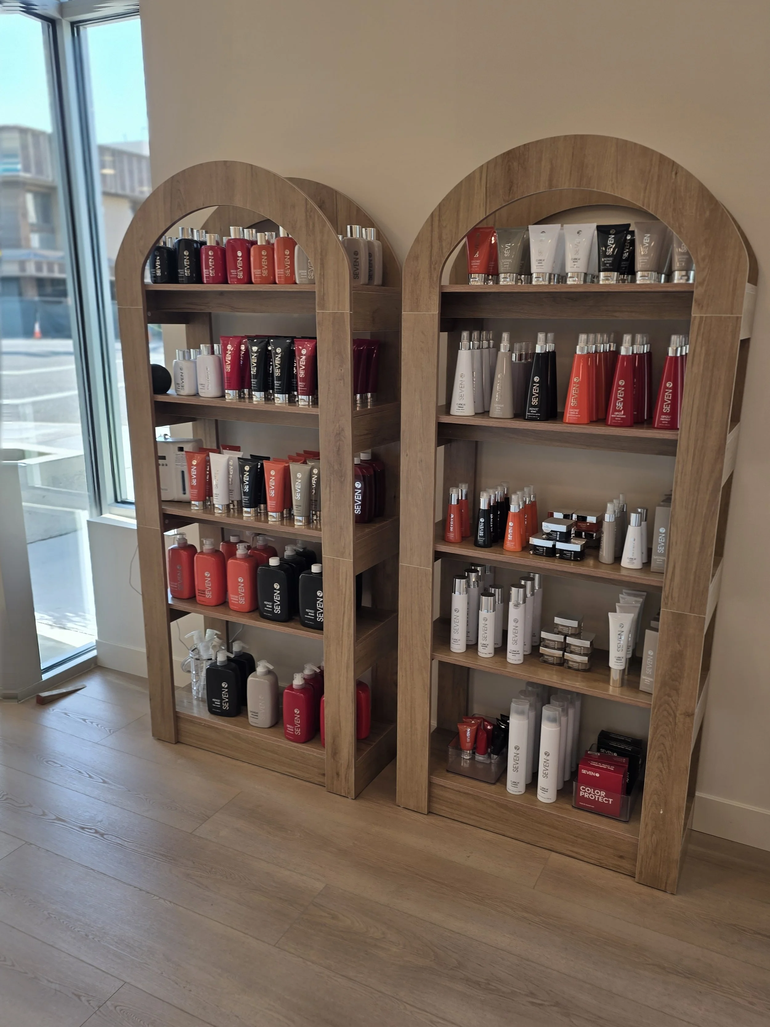 Two wooden display shelves filled with various hair and skincare products, situated near a large window with a cityscape view outside.