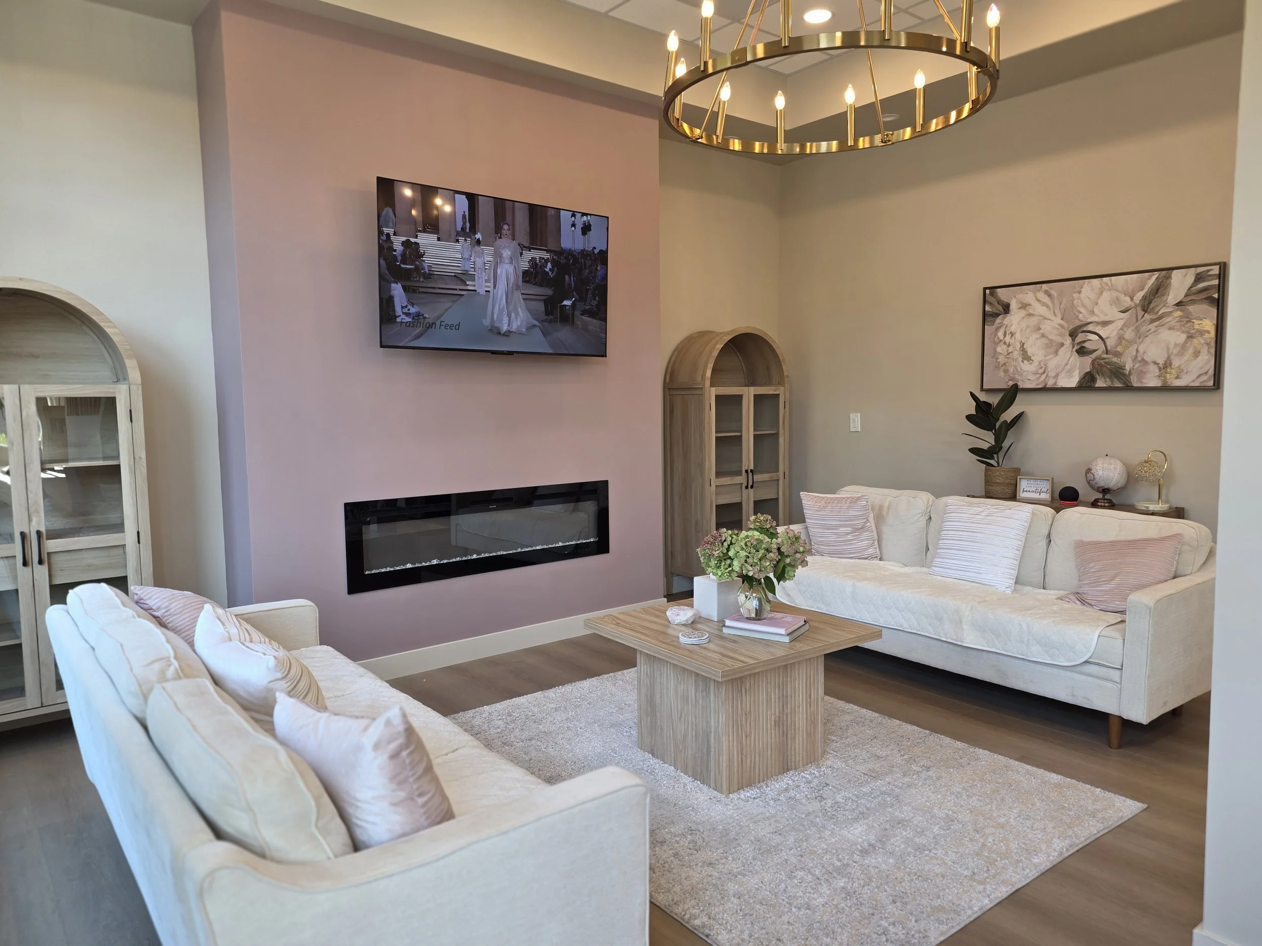 Living room with two white sofas, a wooden coffee table with a bouquet of flowers, a wall-mounted TV, a fireplace, and wall art, with a chandelier hanging from the ceiling.