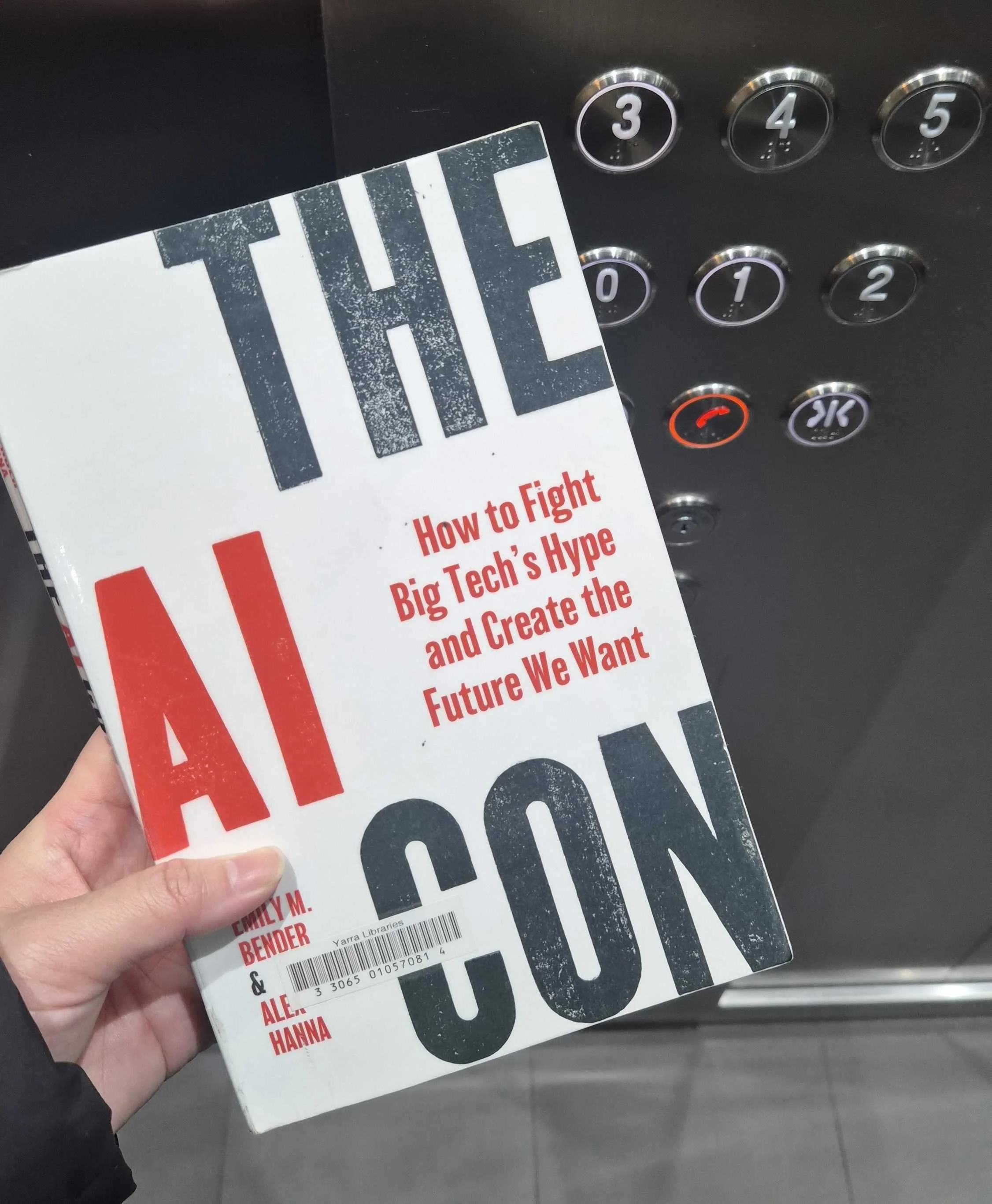 A woman's hand holding up the book "The AI Con" by Emily M. Bender & Alex Hanna in a lift. The book has a white cover with red and black text.