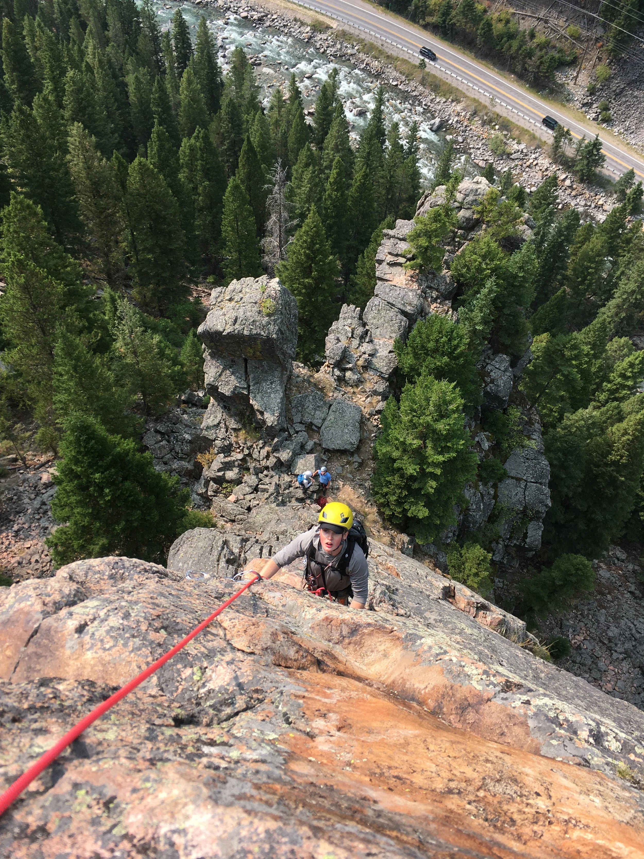 A person wearing a yellow helmet and climbing gear is ascending a rocky cliff. Two other climbers are visible further down the rock face. In the background, there is a forest of green trees, a river, and a road with cars.