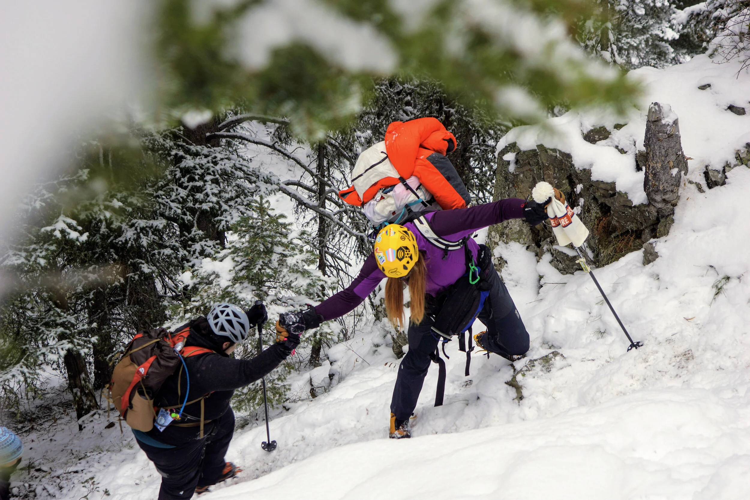 Two hikers assisting each other up a snowy mountain trail in a winter forest, wearing helmets and backpacks.