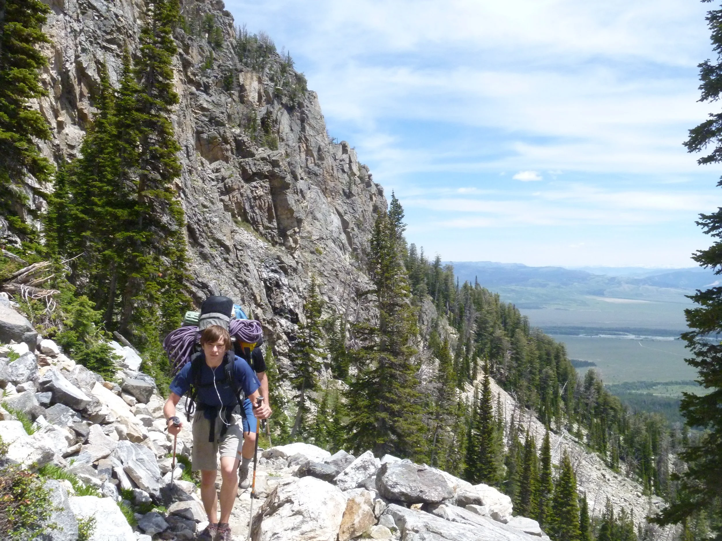 Young boy hiking on rocky mountain trail with backpack and walking sticks in a forested mountain landscape.