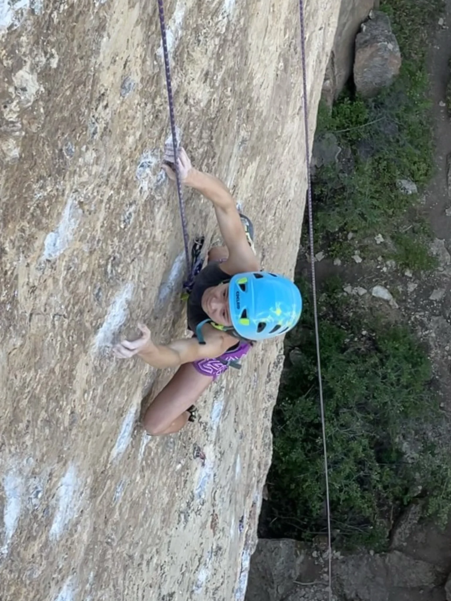 A woman rock climbing on a steep outdoor rock face, wearing a blue helmet, black top, purple shorts, and climbing shoes.