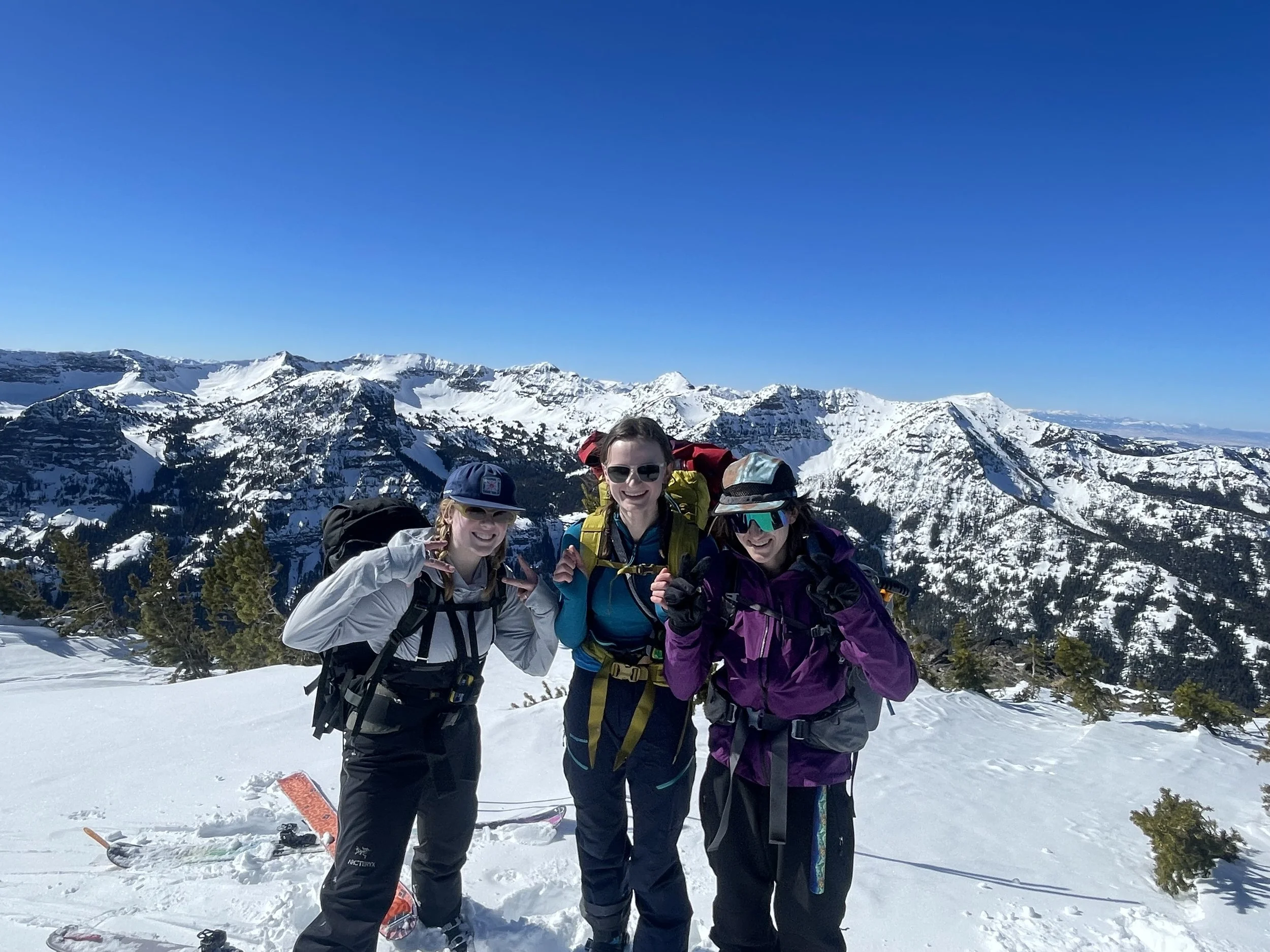 Three women in winter gear smiling and posing on a snowy mountain with snow-covered peaks and a clear blue sky in the background.