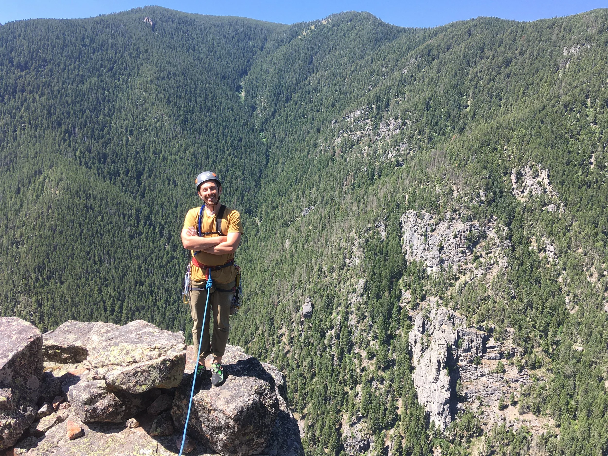 A man wearing a helmet and climbing gear standing on a rocky ledge with a forested mountain range in the background.