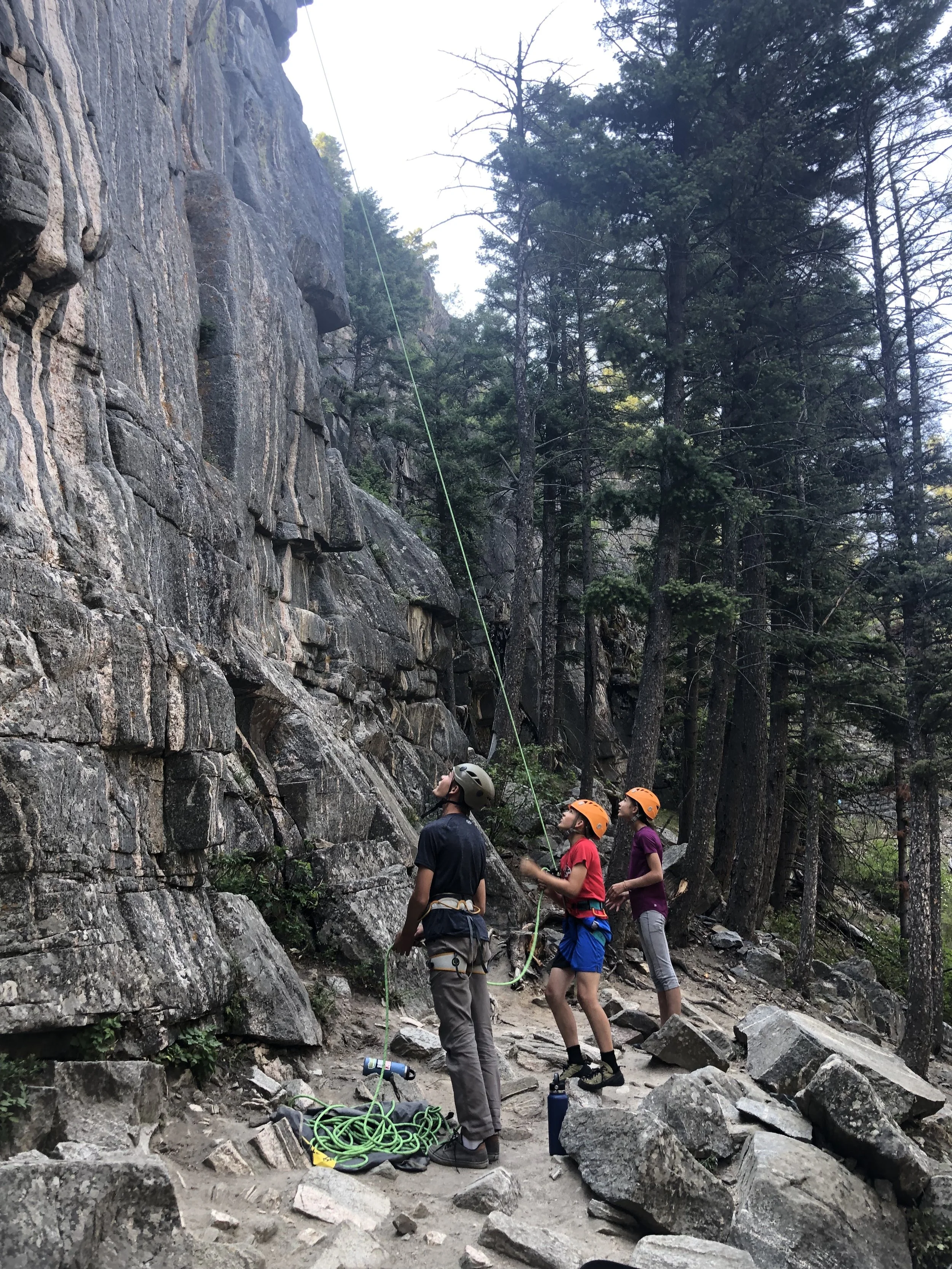 Three people in helmets preparing to climb a rock wall in a forested outdoor area.