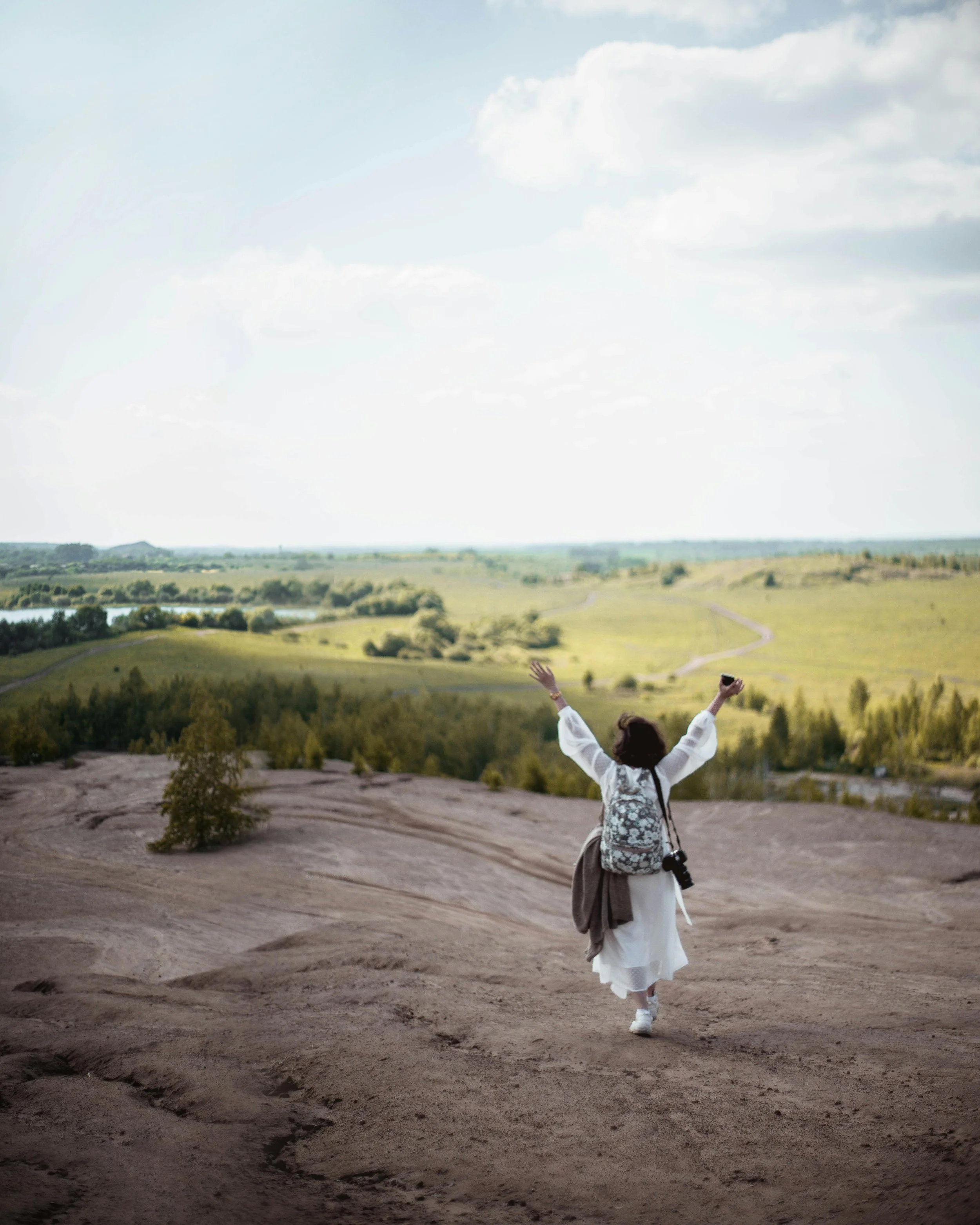 Person with a backpack walking on a dirt path in an open landscape with green hills, trees, and a partly cloudy sky.