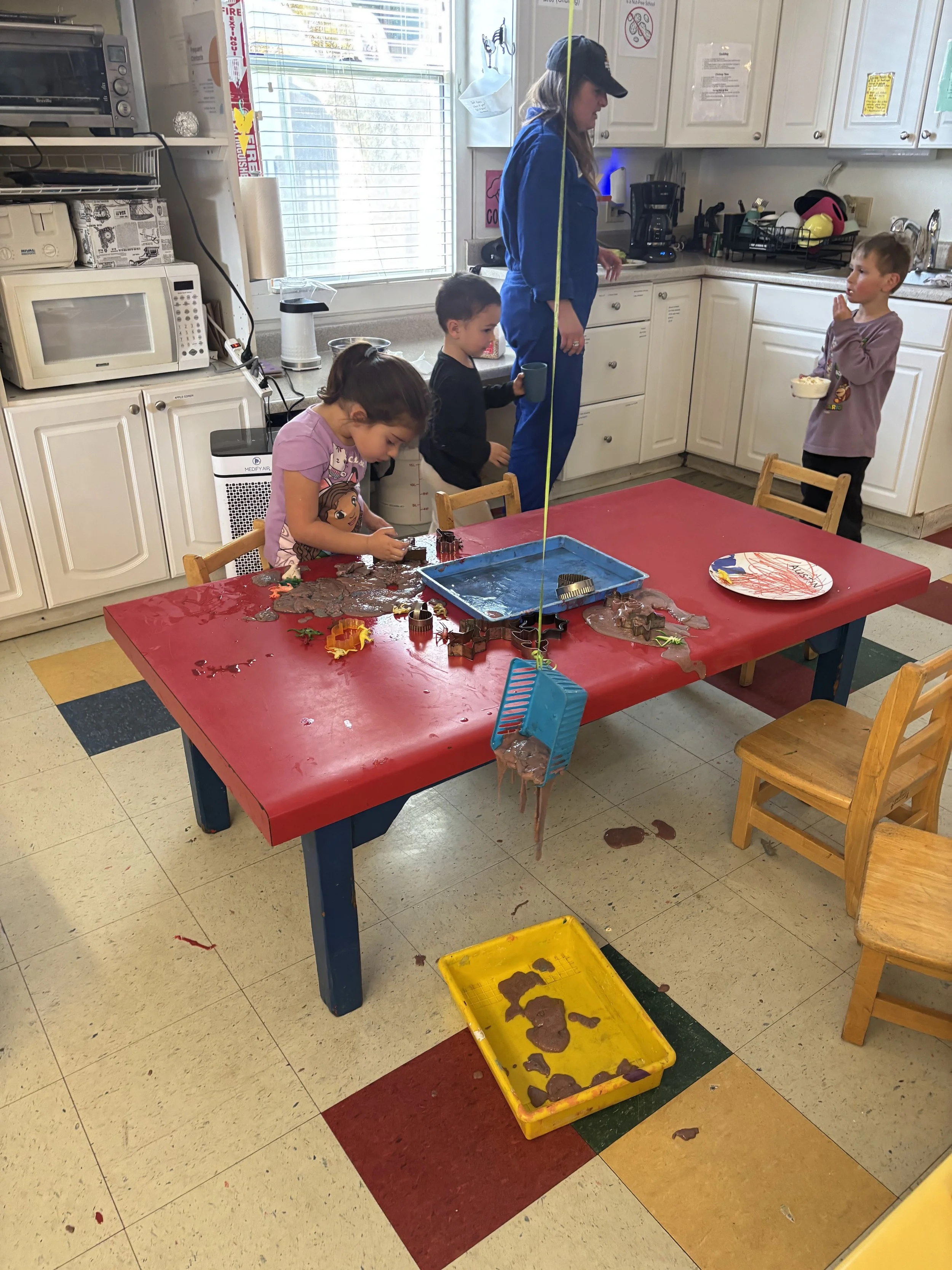 A teacher observes kids making brown goo that looks like slime - they are making a dinosaur playscape with cookie cutters, dinosaurs, and insects.