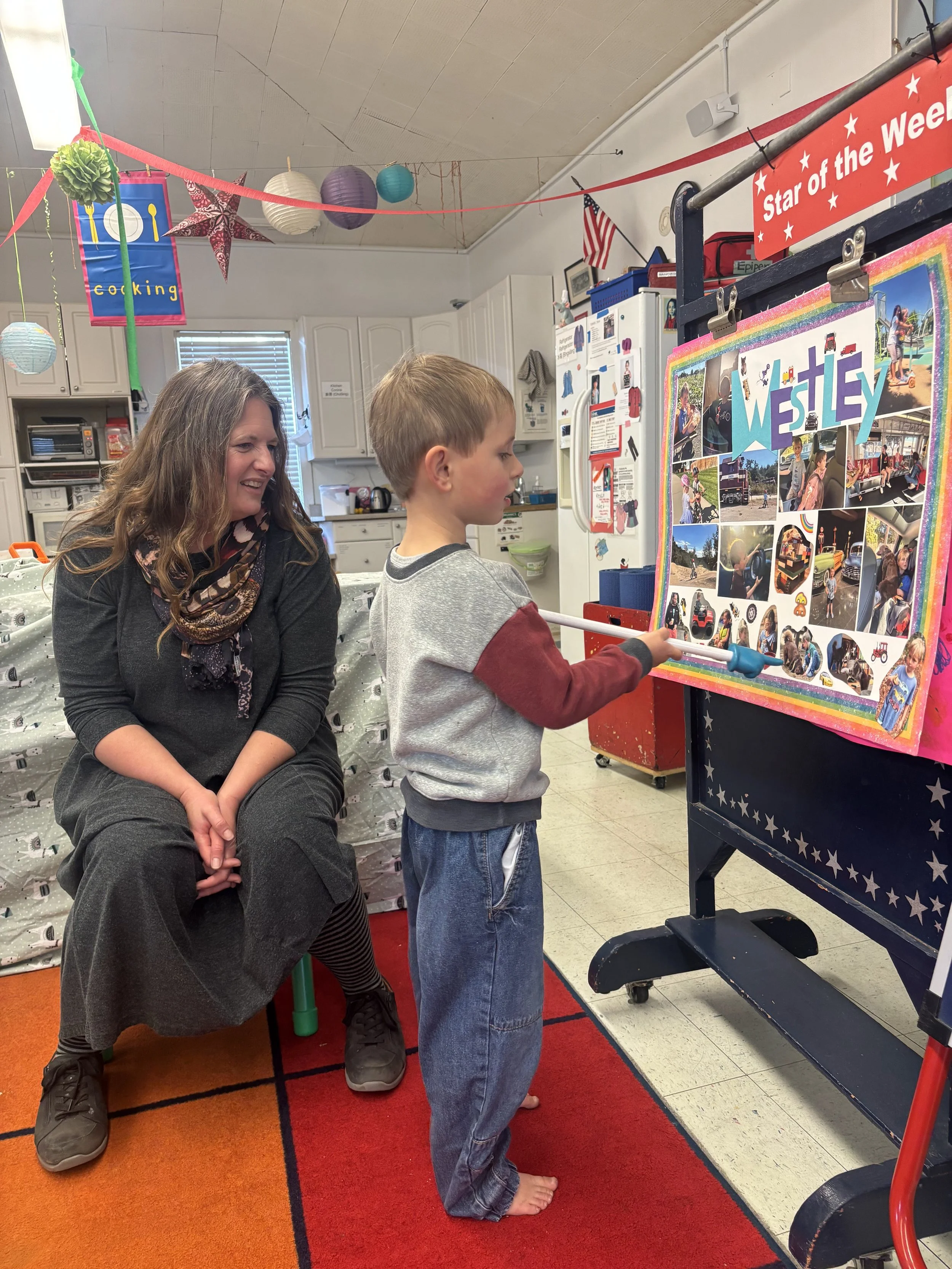 A teacher watches a child present their 'Star of the Week' poster where they call on classmates to select pictures which they then talk about and describe in detail after answering questions from the teacher.