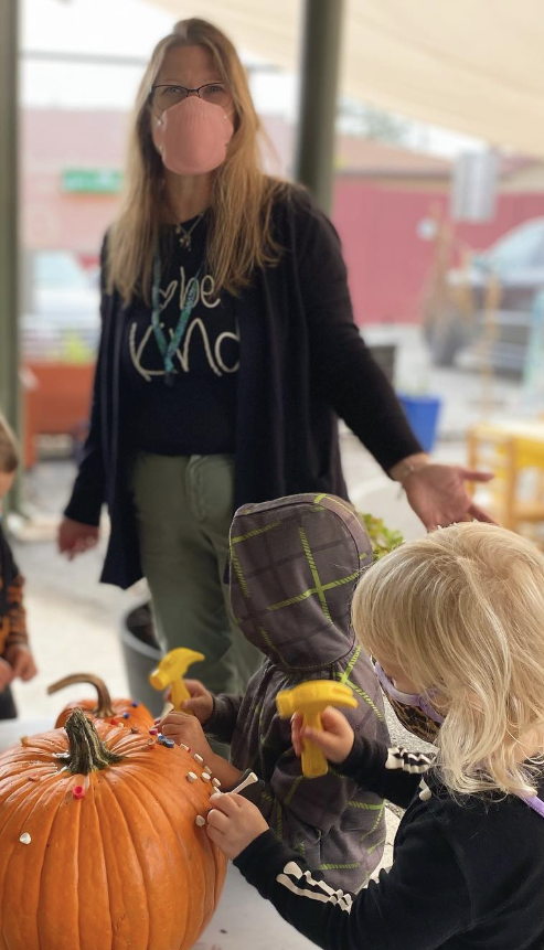 A teacher wearing a facemask observes students hammering golf tees into pumpkins.