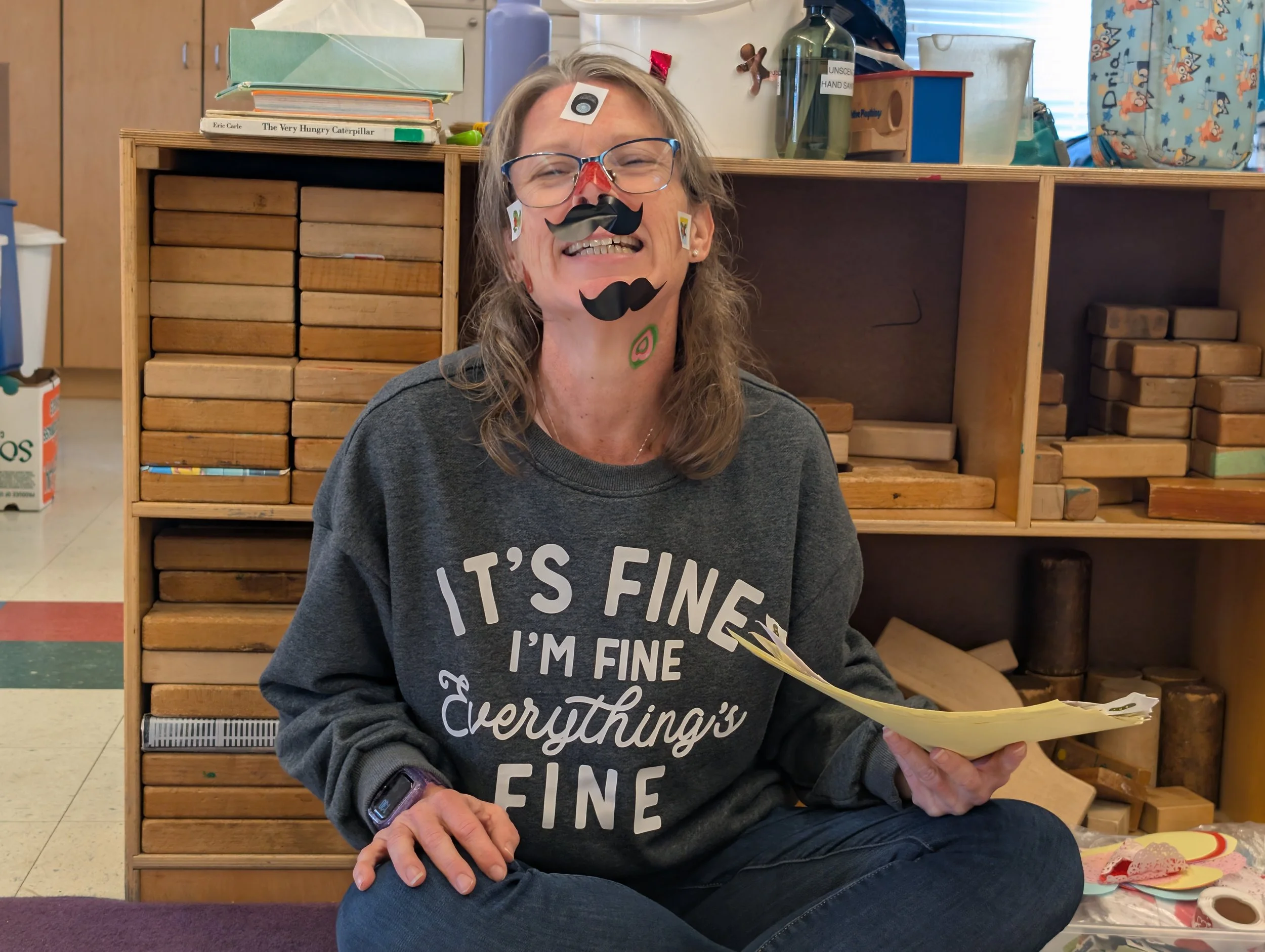 A teacher sits in front of a shelf holding wooden blocks with stickers all over her face.