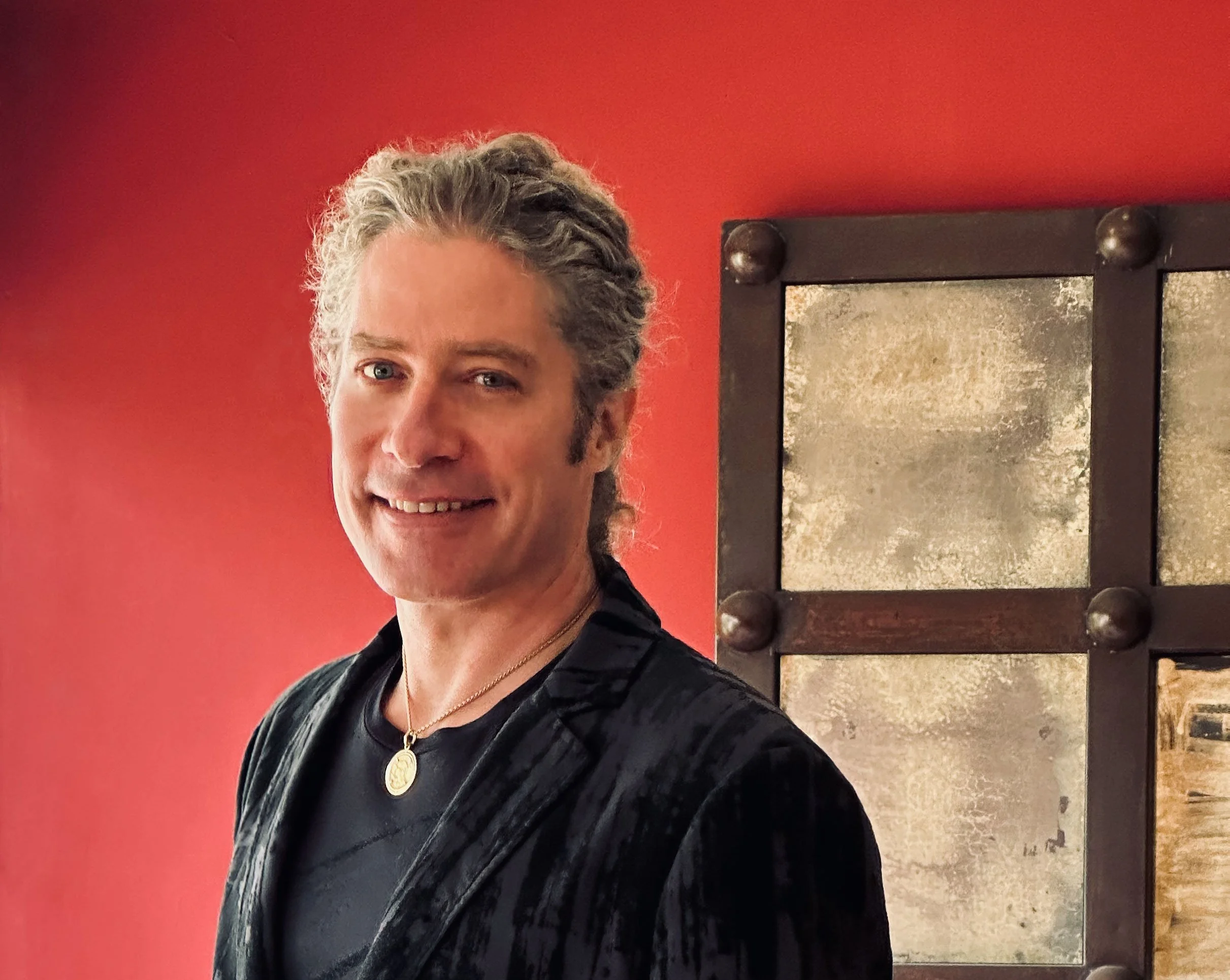 Close-up of a smiling woman with gray hair, wearing a black leather jacket and a gold necklace, standing in front of a red wall and a black-framed window.