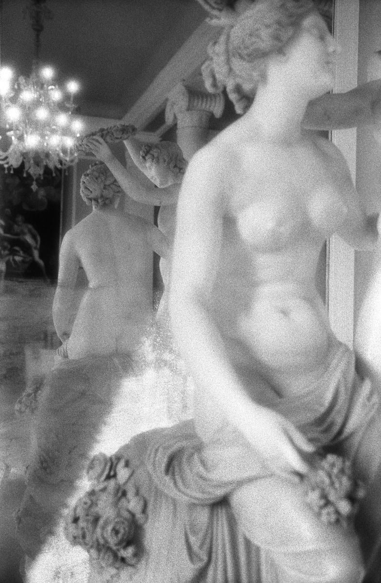 Black and white photograph of classical-style marble statues of women in an interior space, with a chandelier visible in the background.