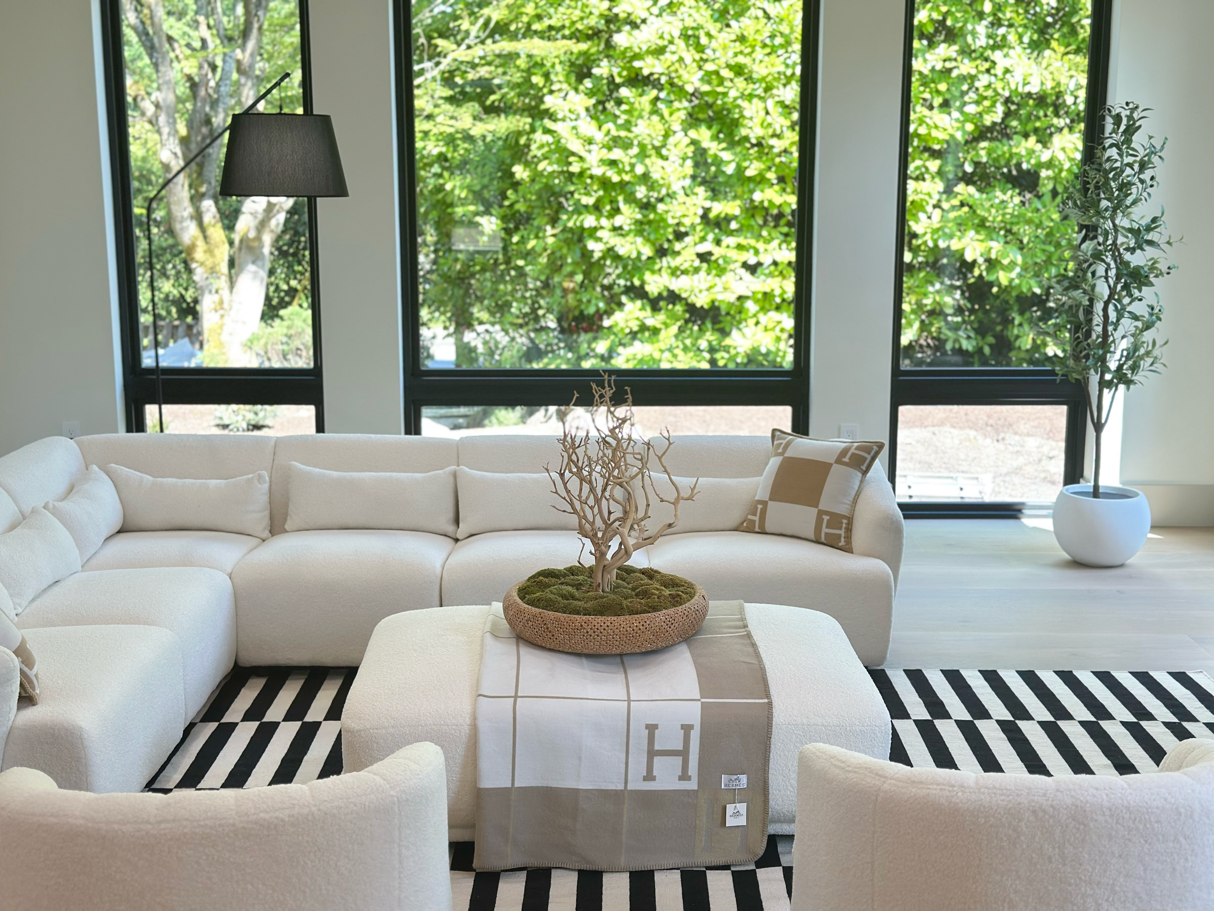 Modern, bright living room with large windows showing green foliage outside, white sectional sofa, a decorative pillow with an 'H' pattern, a black-and-white striped rug, a center ottoman with a decorative bowl holding a small tree, and a tall potted