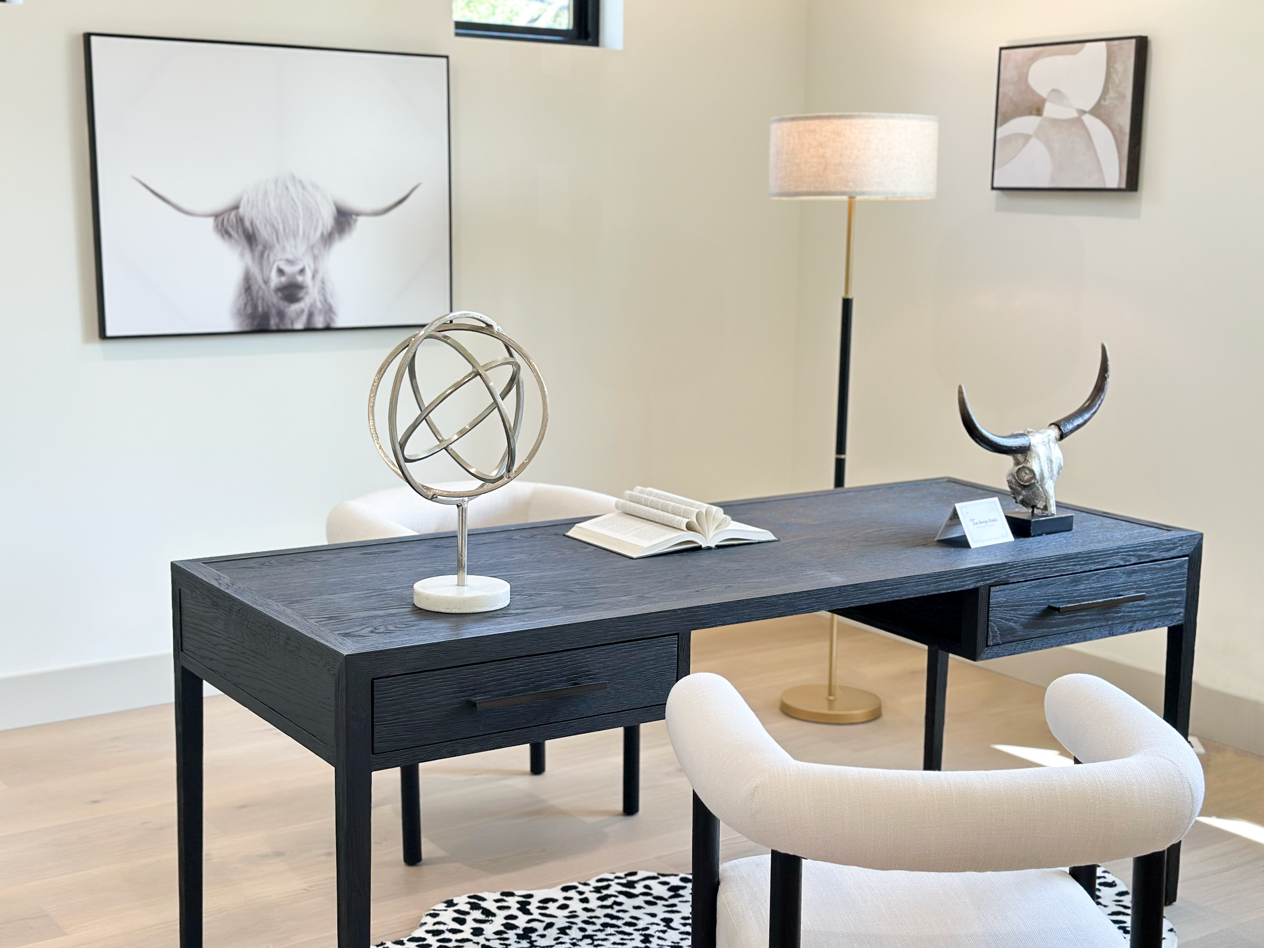 Modern office or living room with a dark wood desk, white armchair, black and white animal print rug, and two framed black-and-white animal photographs on the wall, including a highland cow skull sculpture and a decorative metal orb sculpture on the 