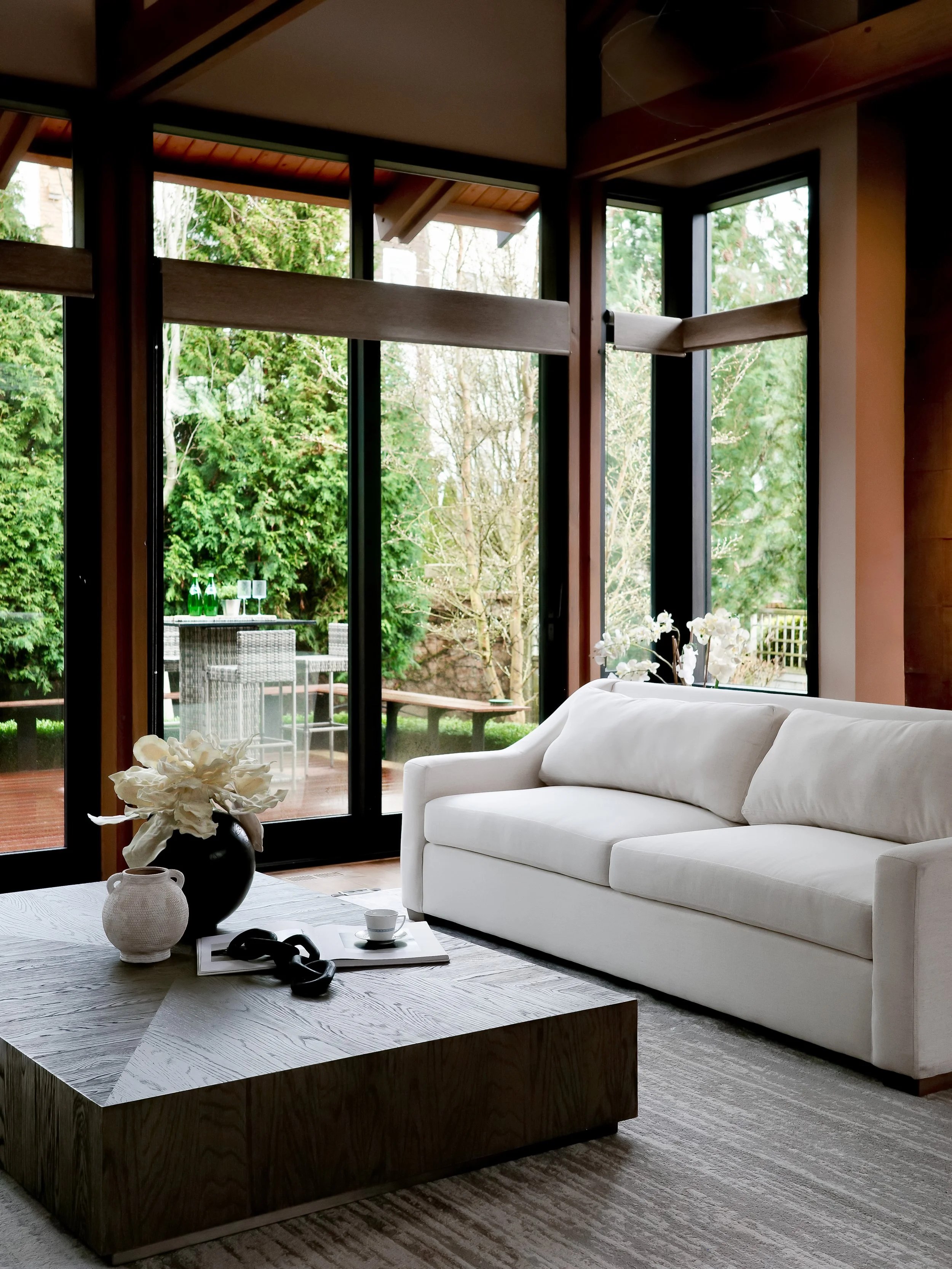 A modern dining room with a white oval table, four wooden chairs with beige cushions, a potted plant centerpiece, and a wall art piece made of dark ceramic vases. There is a window bringing in natural light.