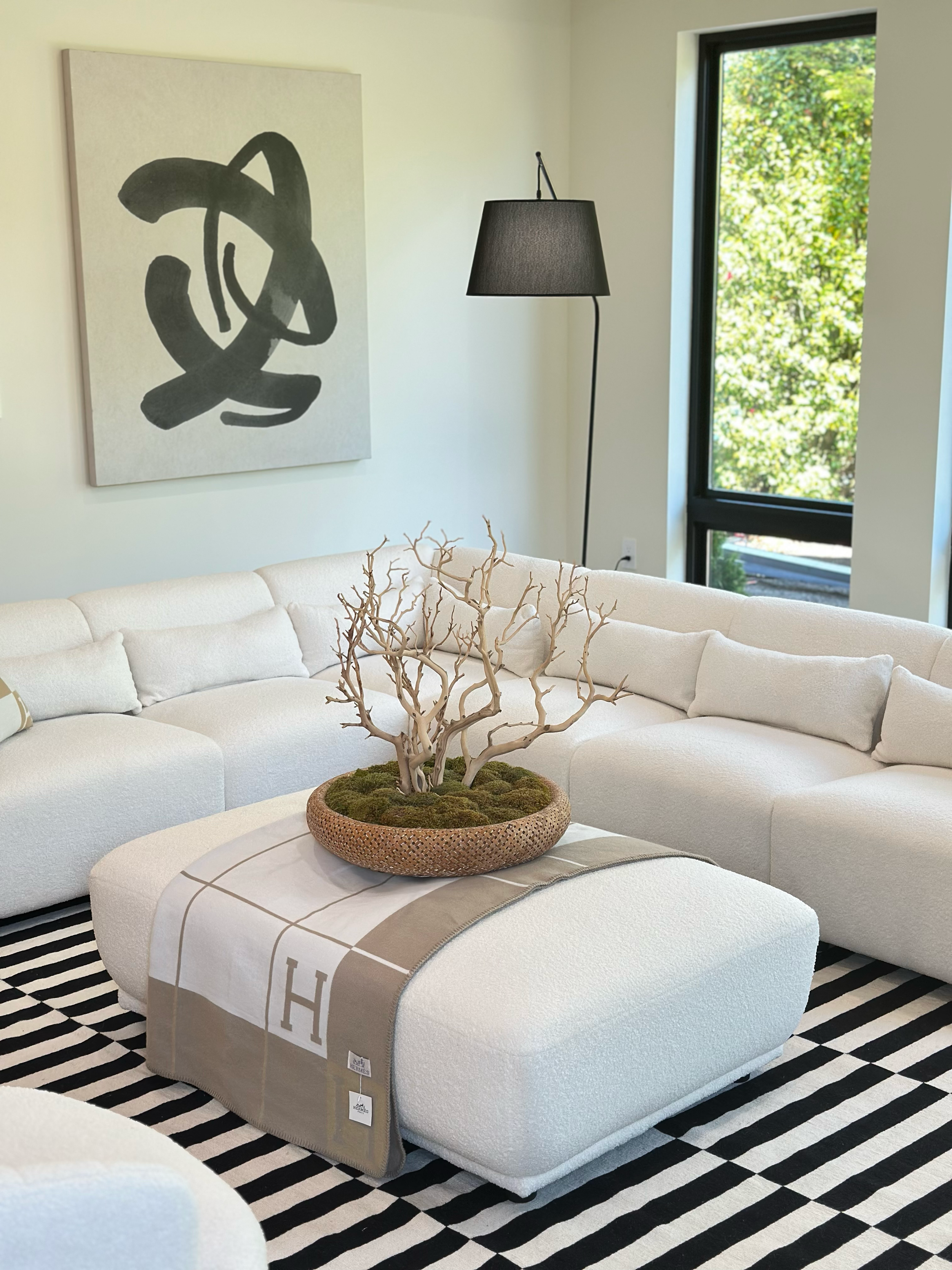 Modern living room with white sectional sofa, decorative branch centerpiece on ottoman, black and white striped rug, black floor lamp, and large window showing greenery outside, with an abstract black and white art piece on the wall.