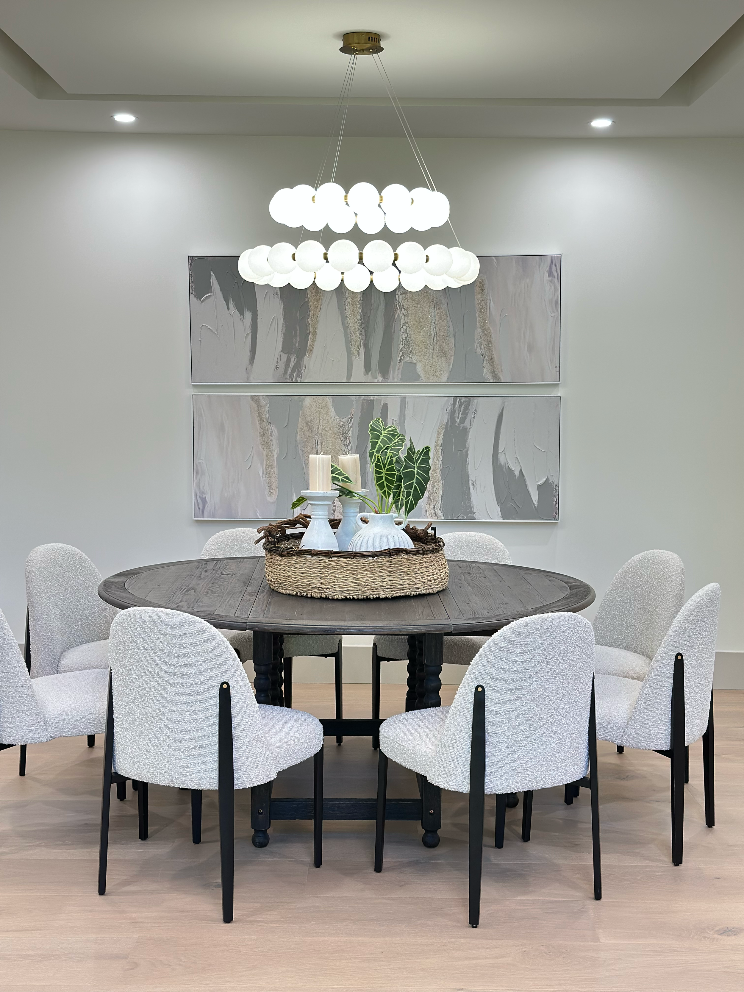Dining room with round wooden table, white upholstered chairs, and modern chandelier with white globes hanging from a ceiling cove. Wall art behind the table featuring two abstract paintings, and decorative centerpiece with plants and candles.