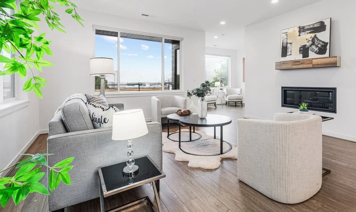 Bright living room with large window, white walls, gray sofa, armchair, coffee table with vase and decor, fireplace, abstract art above fireplace, hardwood floors, and potted plants.