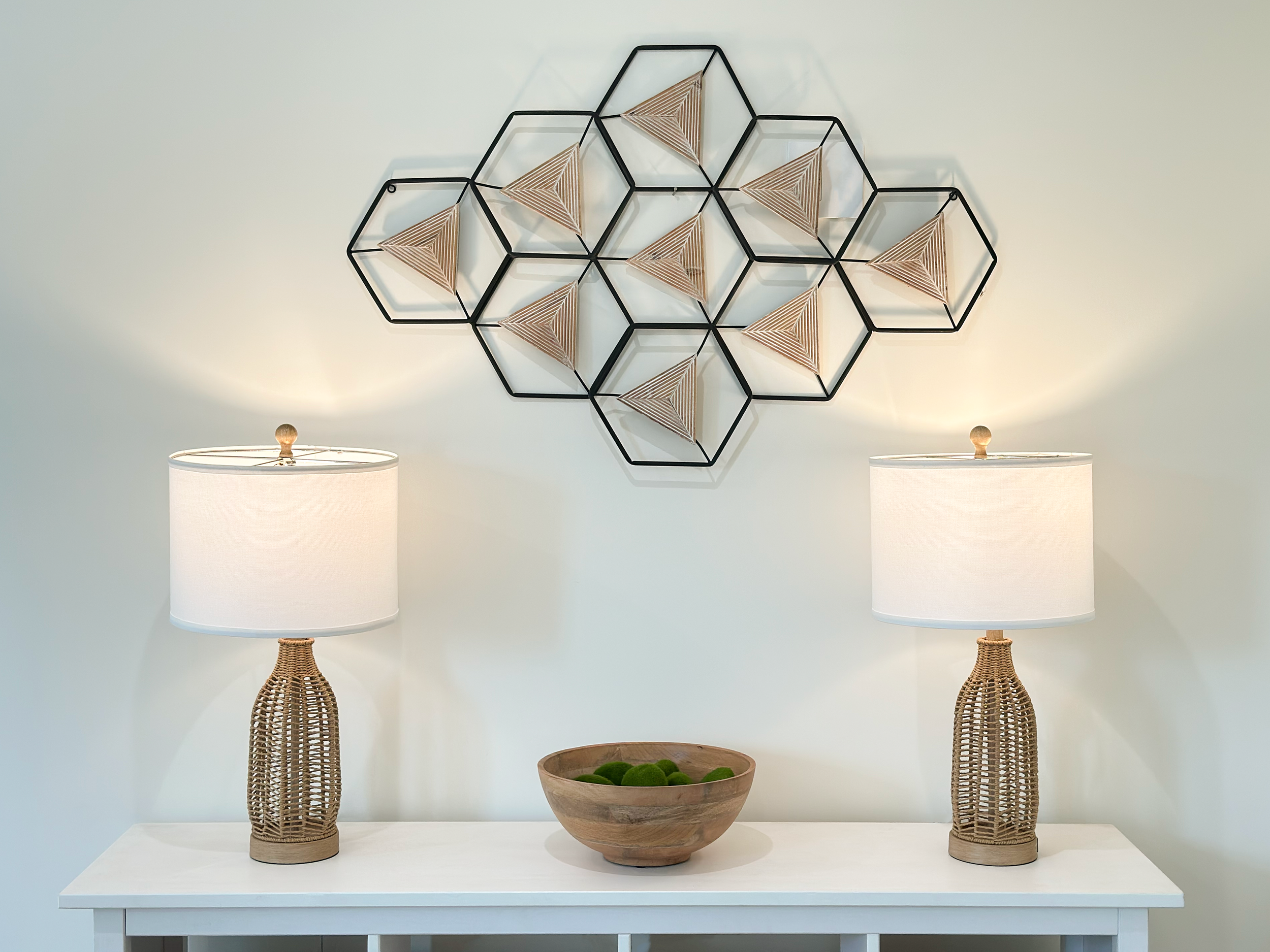 A white console table with two wicker table lamps, a wooden bowl with green moss inside, a wall decoration made of hexagonal black frames with woven geometric designs, on a plain white wall.